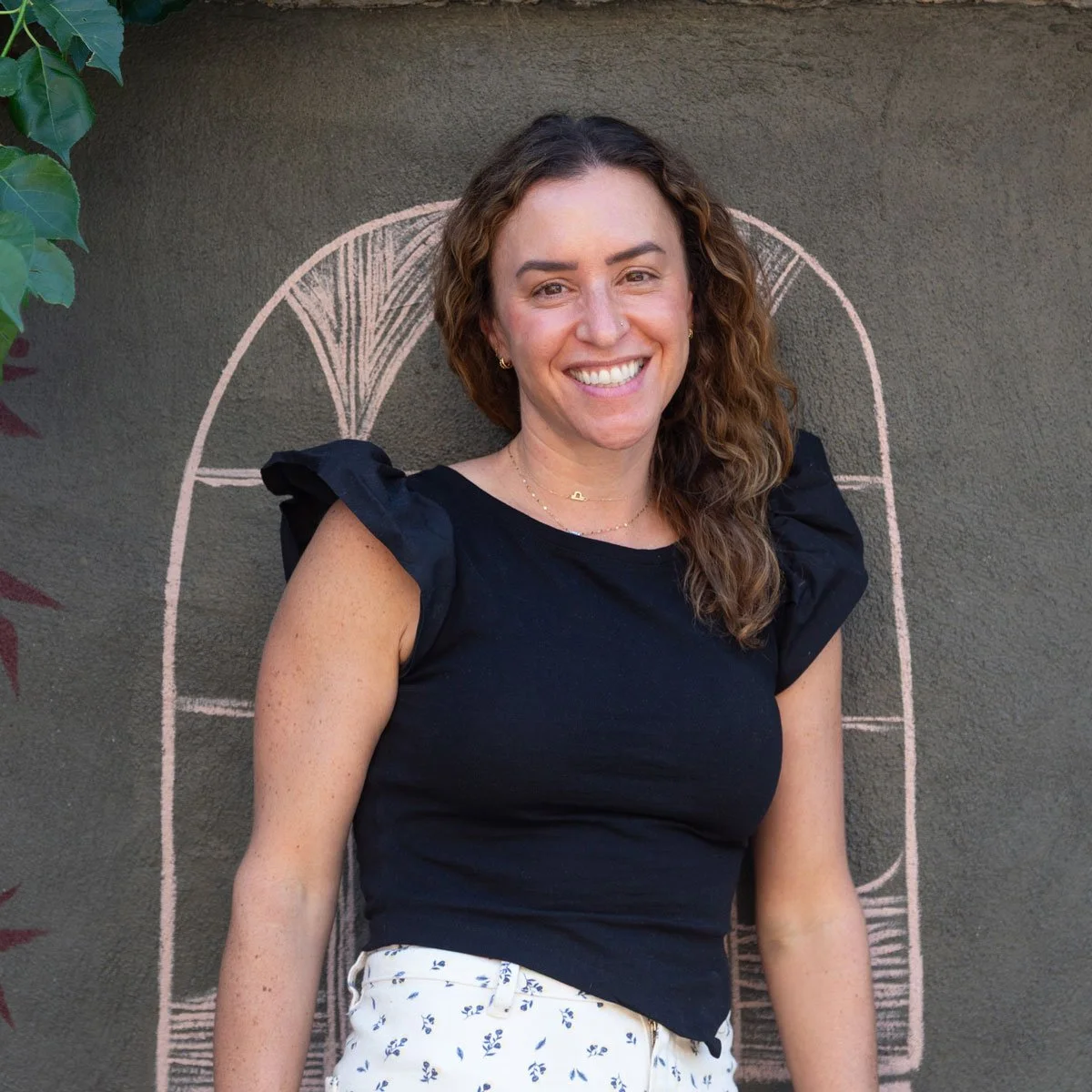 A woman smiling outdoors, standing in front of a green wall with a pink chalk drawing of an arched window in East Falls, Philadelphia.