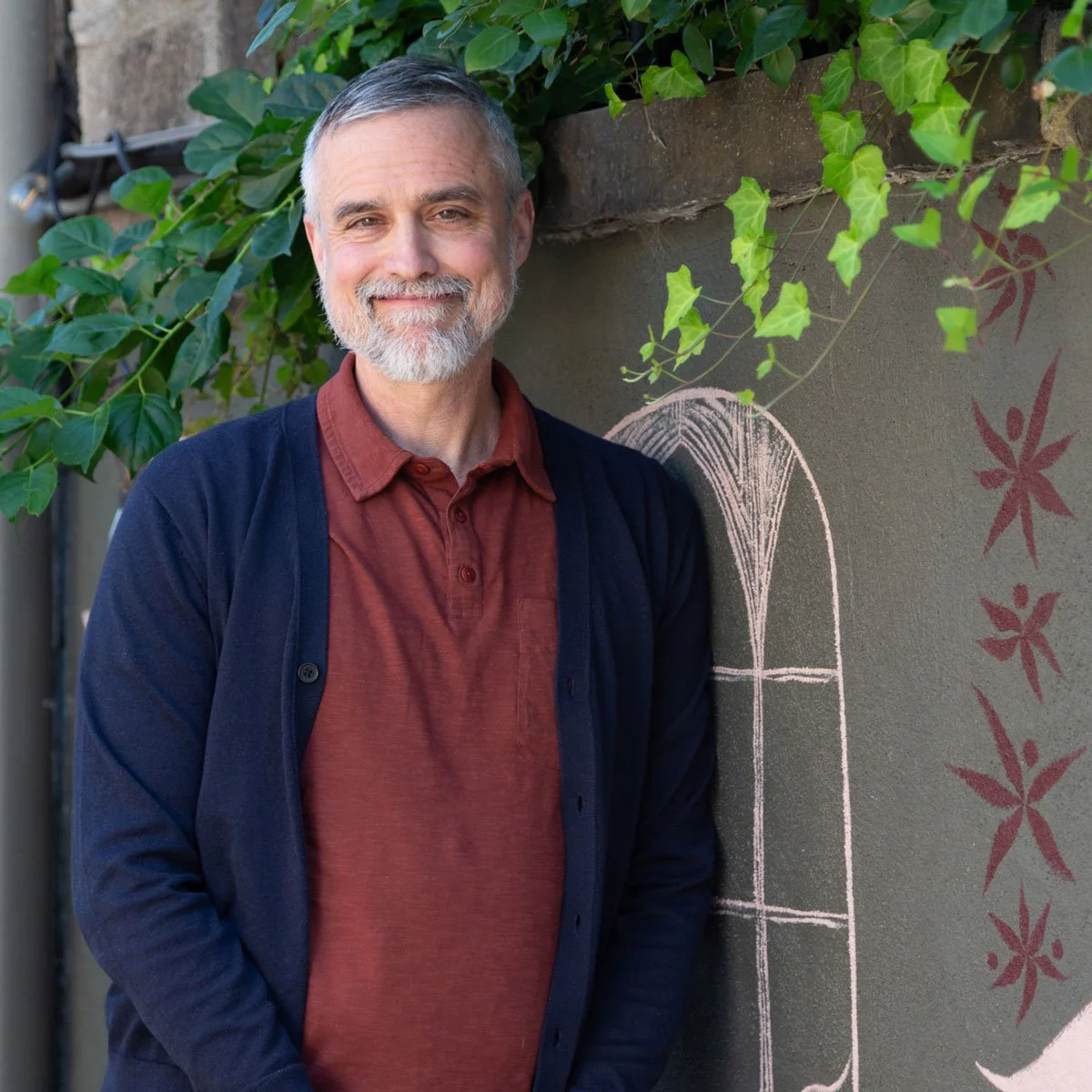 A middle-aged man with gray hair and a beard, wearing a maroon shirt and navy cardigan, smiling while standing outdoors next to a wall with chalk art of a window and red floral patterns, surrounded by green leaves in East Falls, Philadelphia.