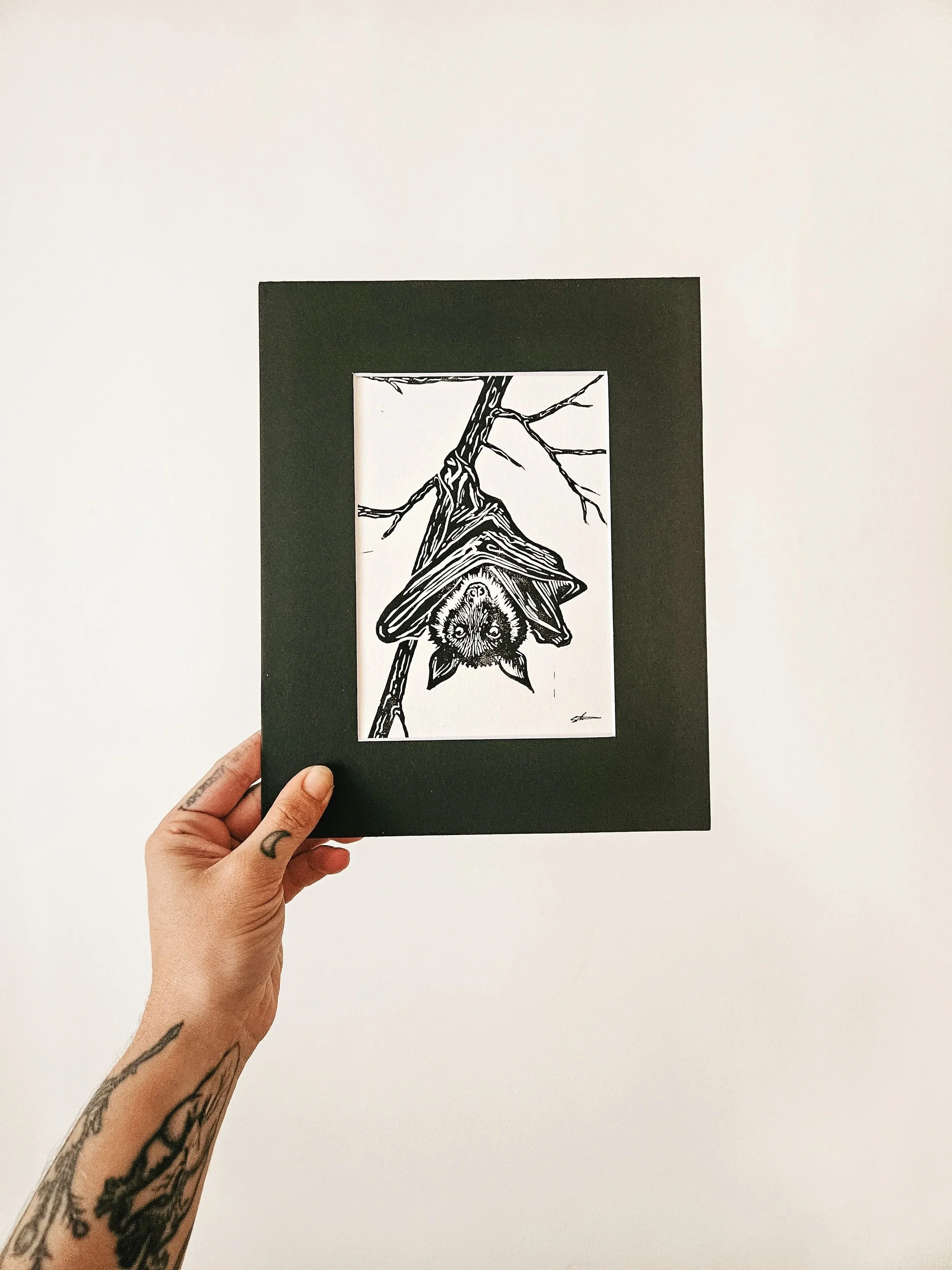 a hand holding up an 8inch by 10 inch hand-pressed linocut print of a bat hanging from a tree branch.