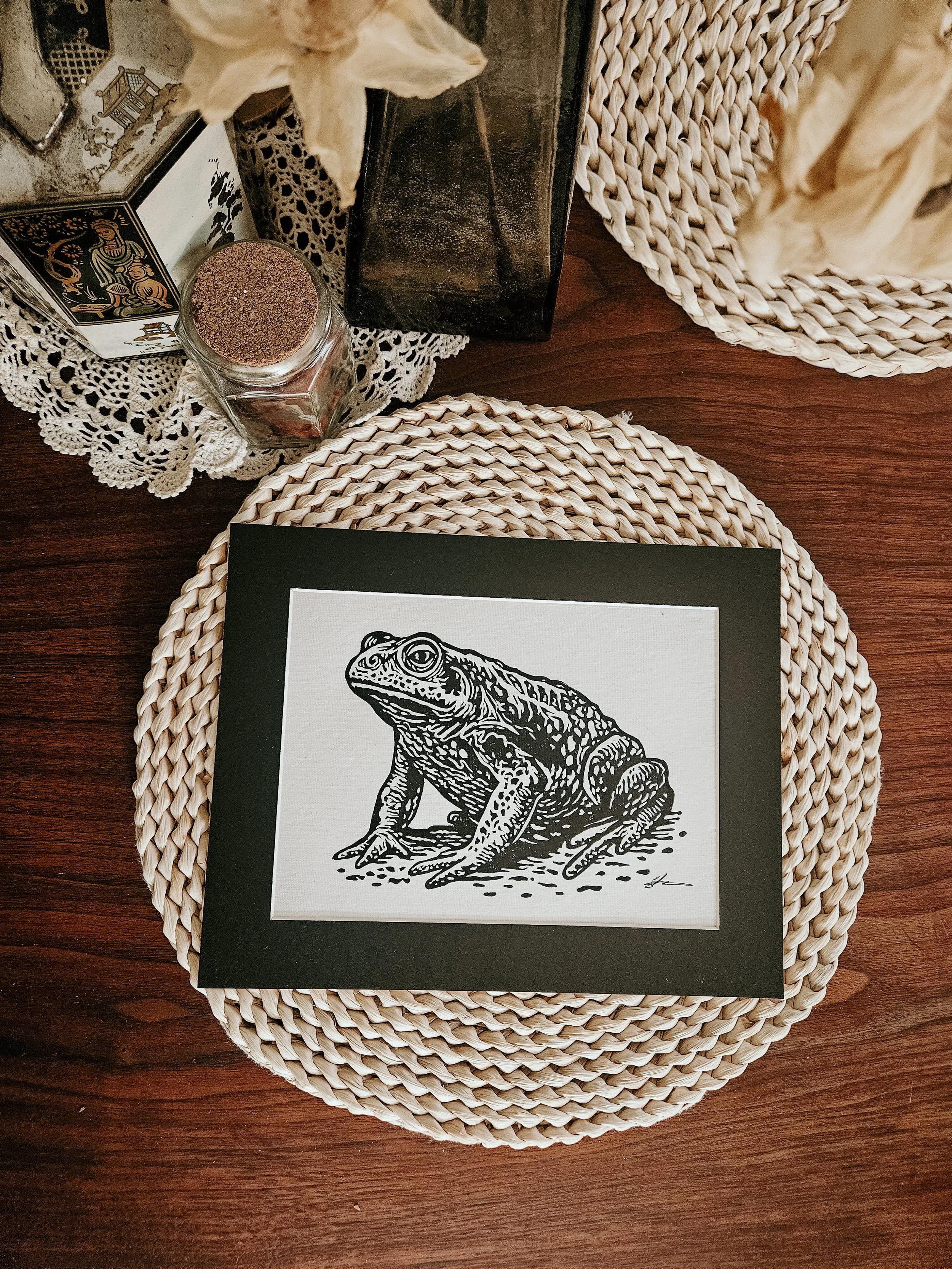 Matted linocut print of a toad by Black Candle Print Co. lying on a placemat.