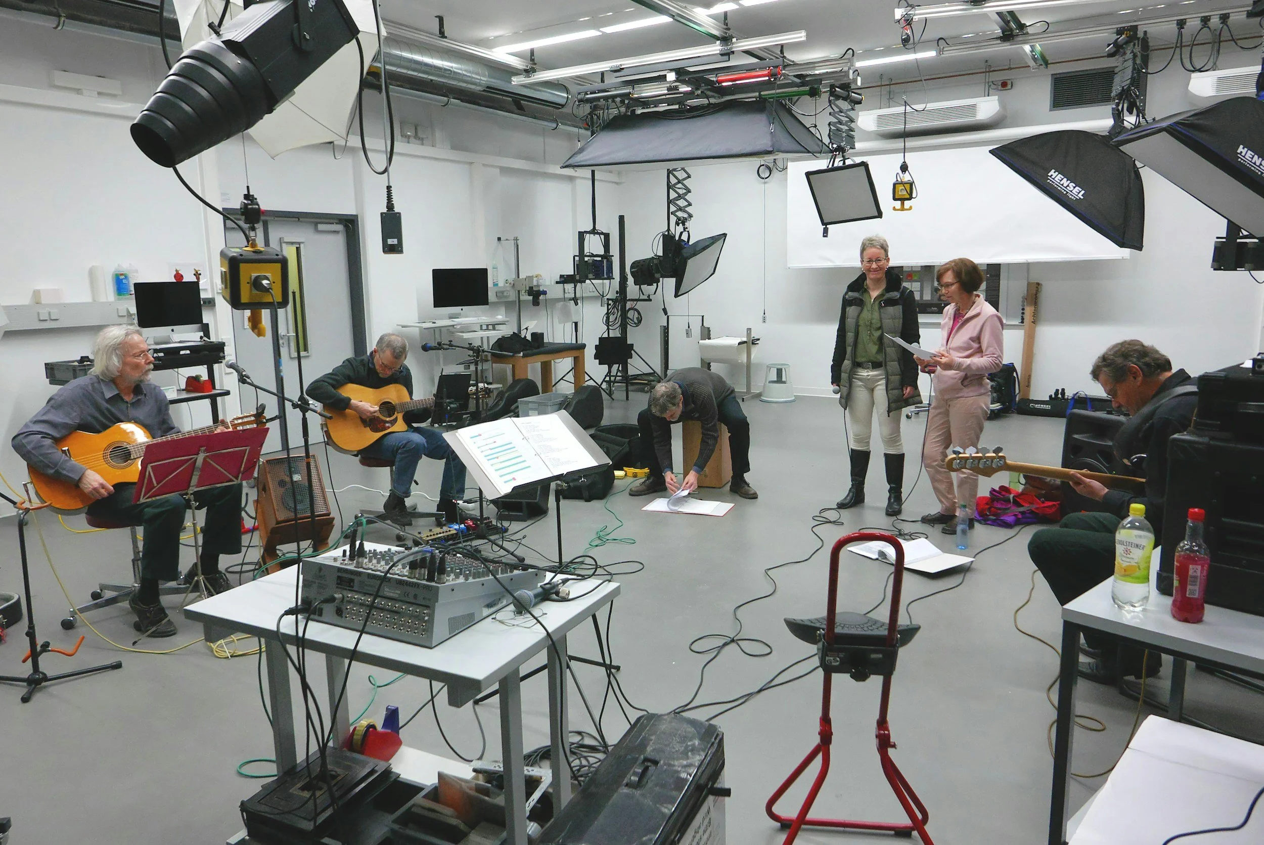 A group of people in a recording studio setup with multiple cameras, lighting equipment, and musical instruments; some are playing guitars and others are preparing or adjusting equipment.