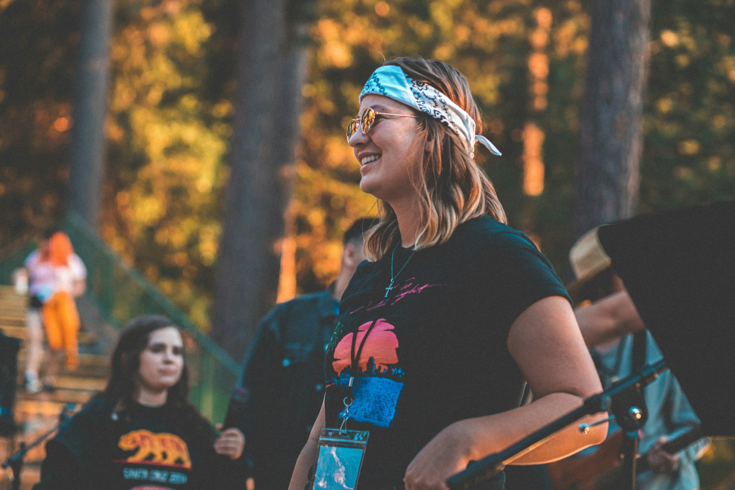 A young woman with curly hair, wearing glasses, a blue bandana, and a black t-shirt with colorful graphics, smiles during an outdoor event in a forest with trees and autumn foliage in the background.