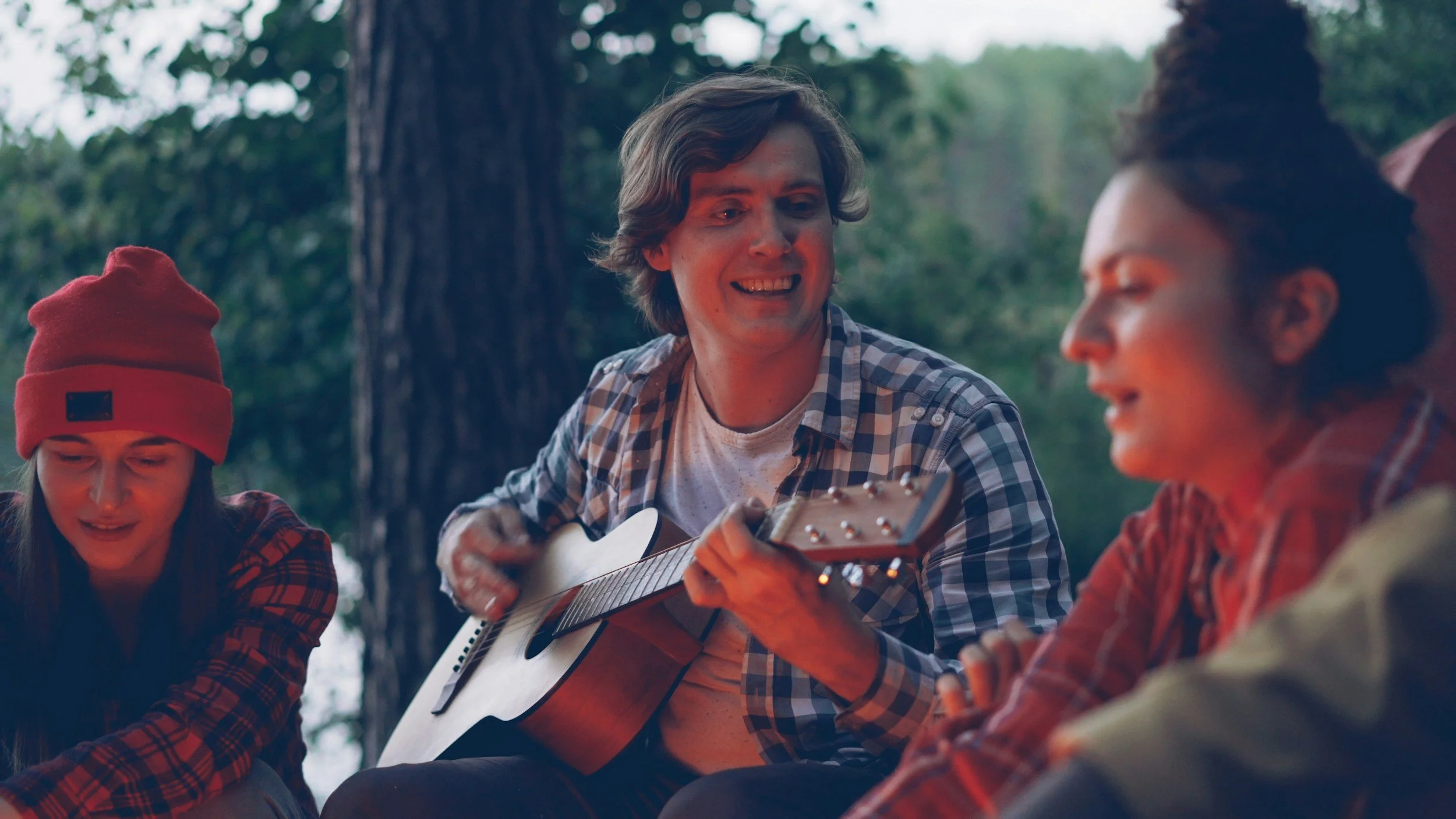 Three young adults sitting outdoors in a forested area, two women and one man, with the man playing guitar and all three smiling and enjoying music together.
