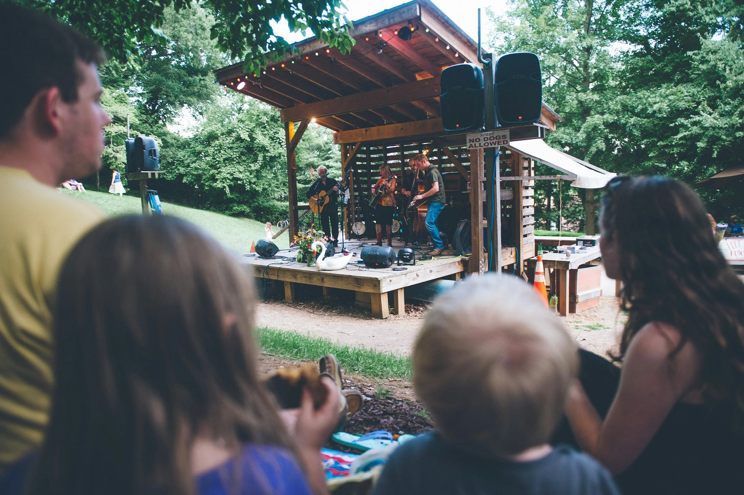A group of people watching a live outdoor music performance on a wooden stage, surrounded by lush green trees, with a sign that reads 'Sorry No Dogs Allowed' and musical instruments being played by band members.