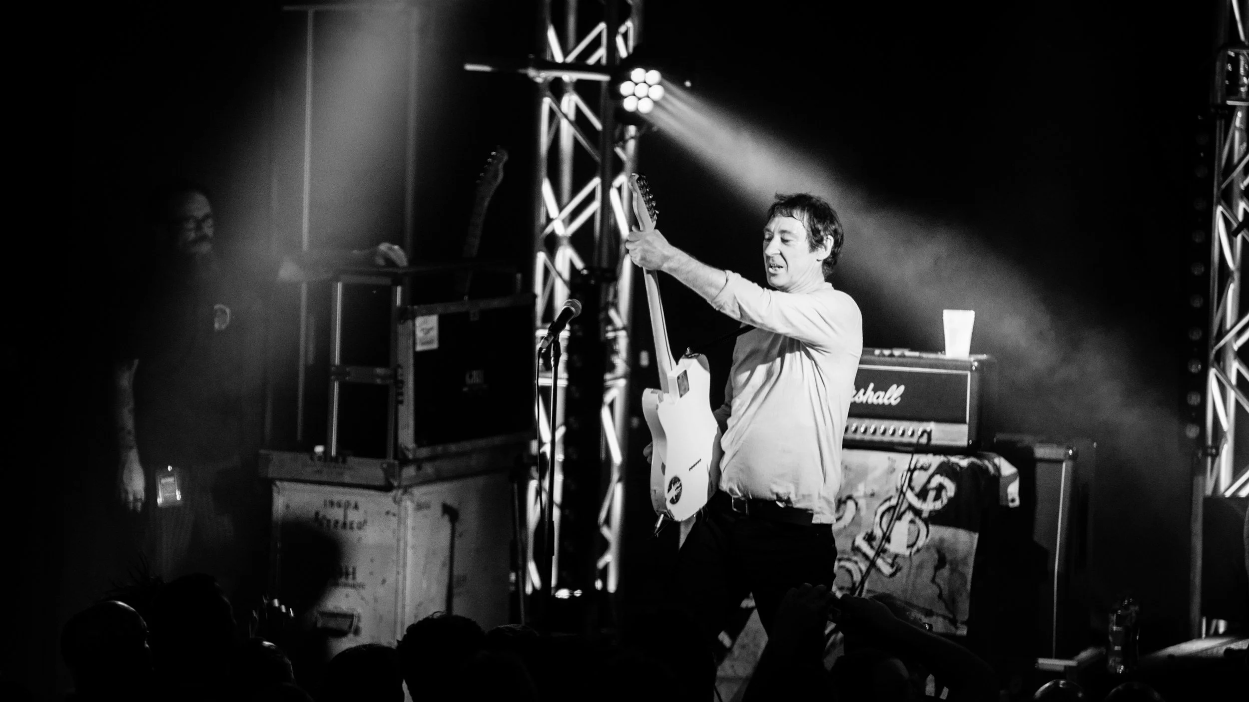A musician with dark hair playing an electric guitar on stage, with stage lighting and equipment, including a Marshall amplifier, visible in the background, during a concert.