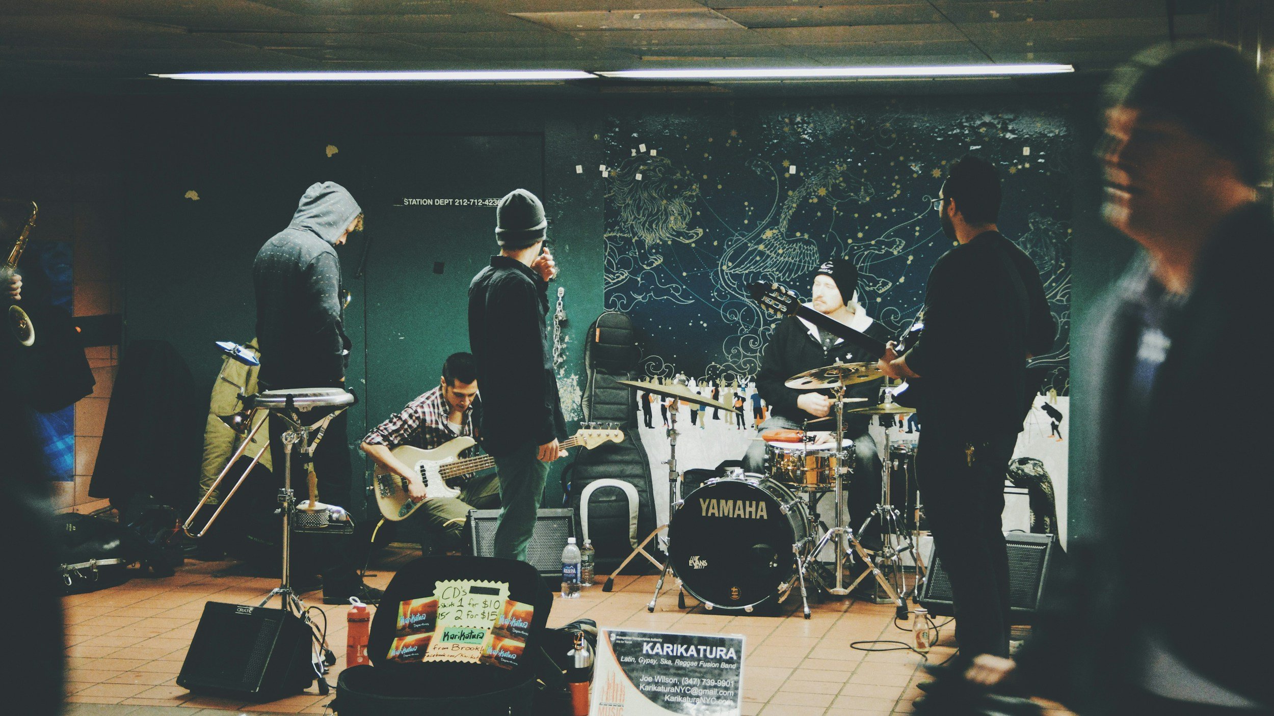 A group of musicians practicing or performing with instruments like bass, drums, and guitar in an indoor space with dark walls and artwork.