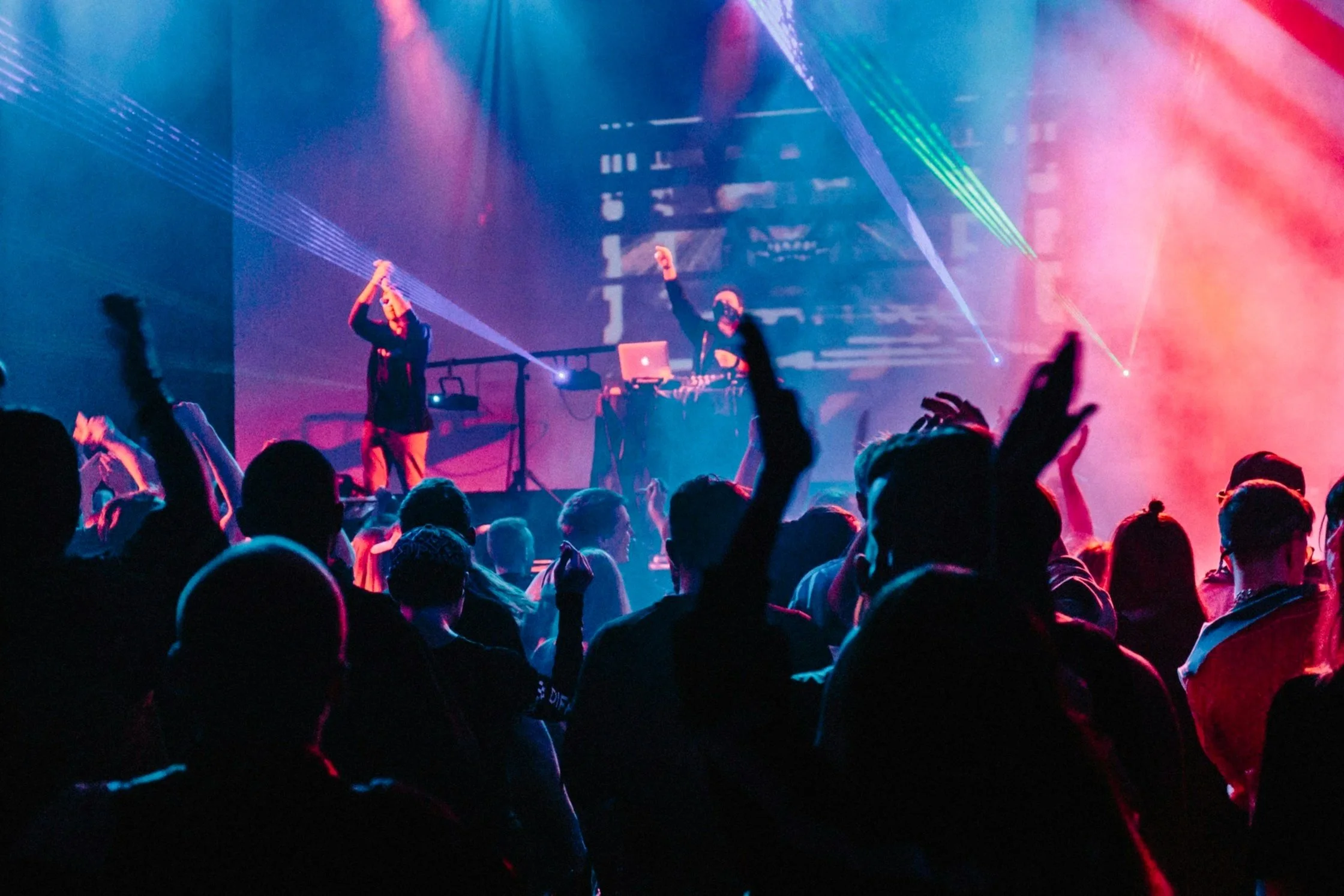 A concert or music event with a crowd of people raising their hands, colorful stage lighting, laser beams, and a DJ or performer on stage with equipment.