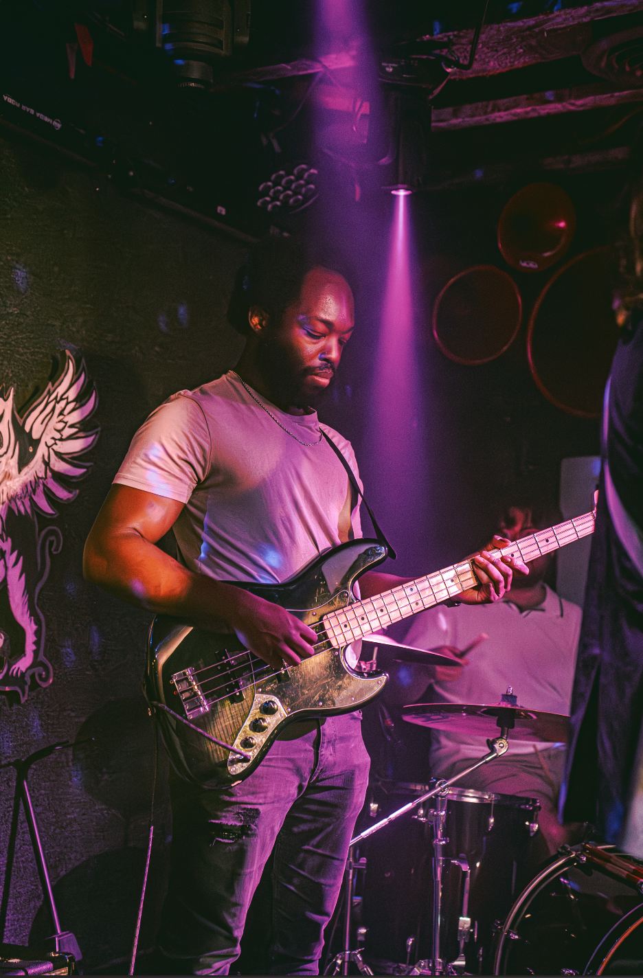 A musician playing an electric bass guitar on stage in a dimly lit venue with purple lighting, a black brick wall, and speakers in the background.