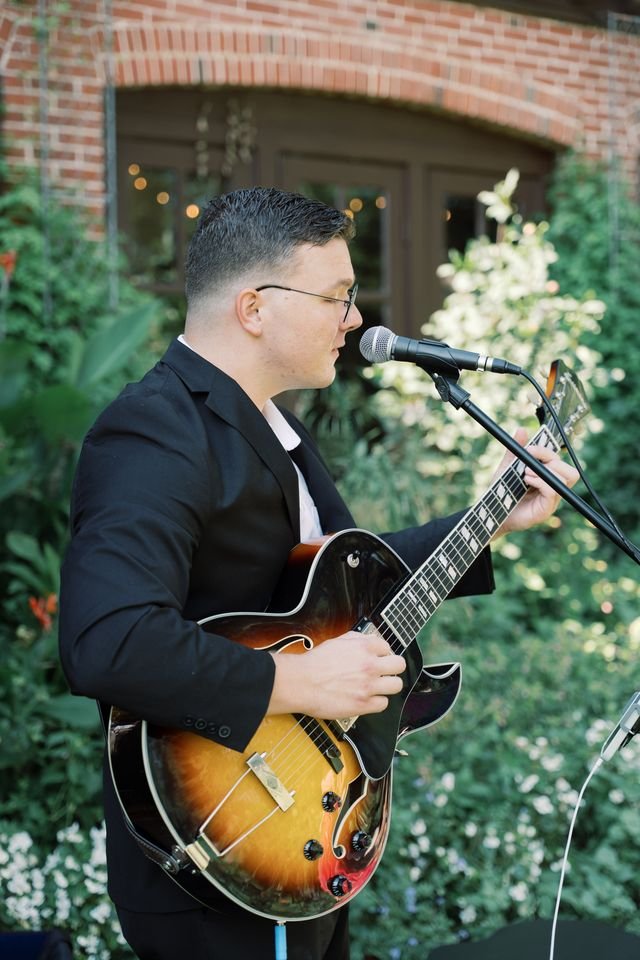 A man wearing glasses and a black suit jacket playing an electric guitar while singing into a microphone outdoors with greenery and a brick building in the background.