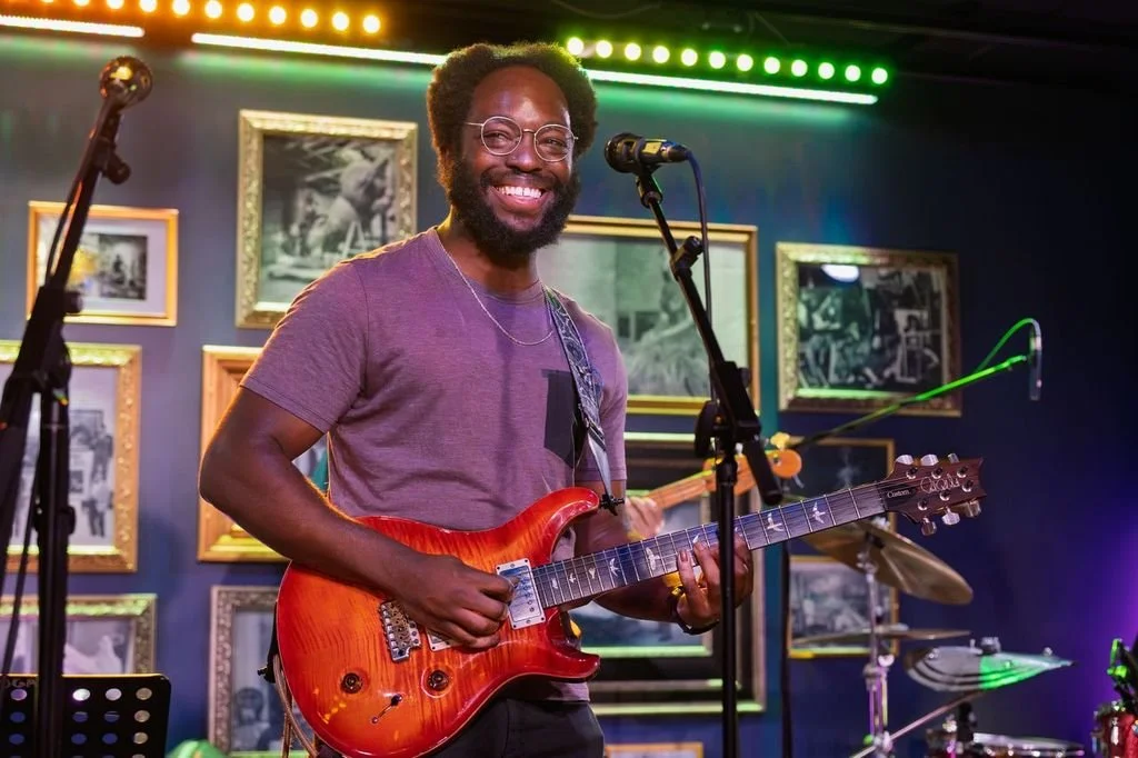 A man smiling while playing an electric guitar on stage, with framed photographs on the wall behind him, colorful stage lighting, and a drum set in the background.