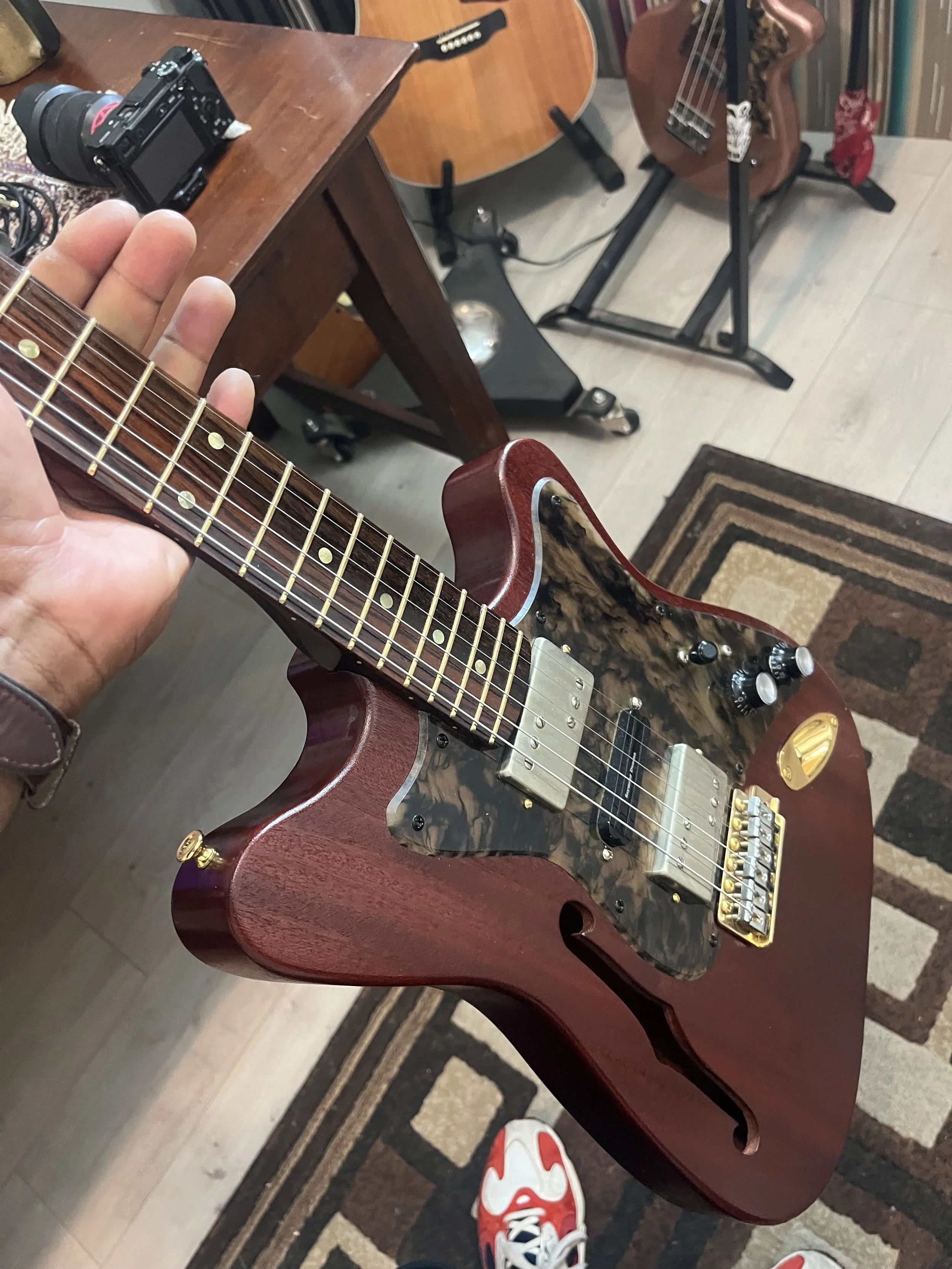 An electric guitar with a smoked tortoise shell pickguard and gold hardware on a wooden table. The guitar has a mahogany body and a suspect design near the neck. A person is wearing red and white sneakers, and the background includes another acoustic guitar, a camera, and various objects on the table.