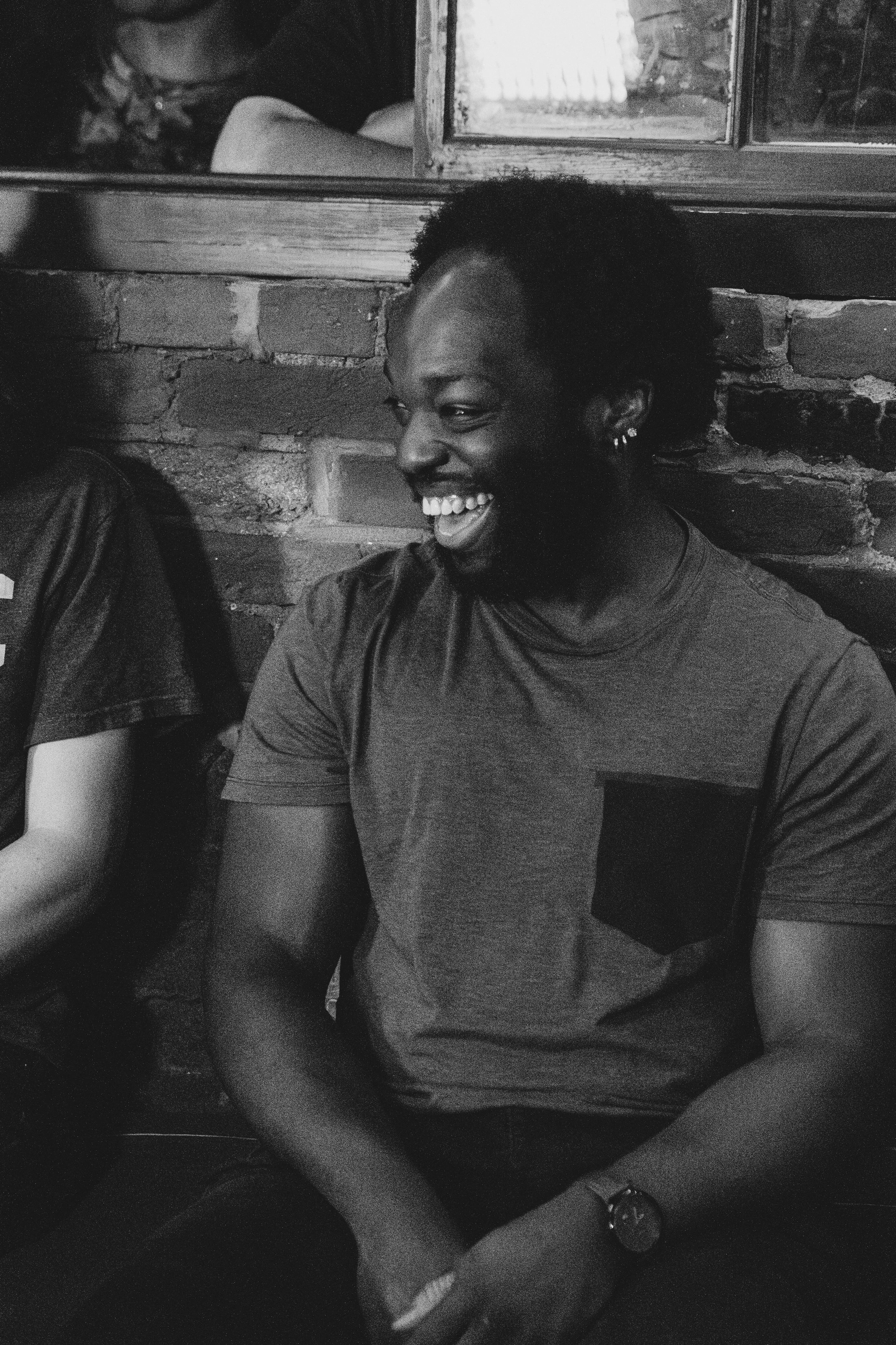 A person smiling, sitting against a brick wall next to a window, in a casual T-shirt, with earrings, in black and white.