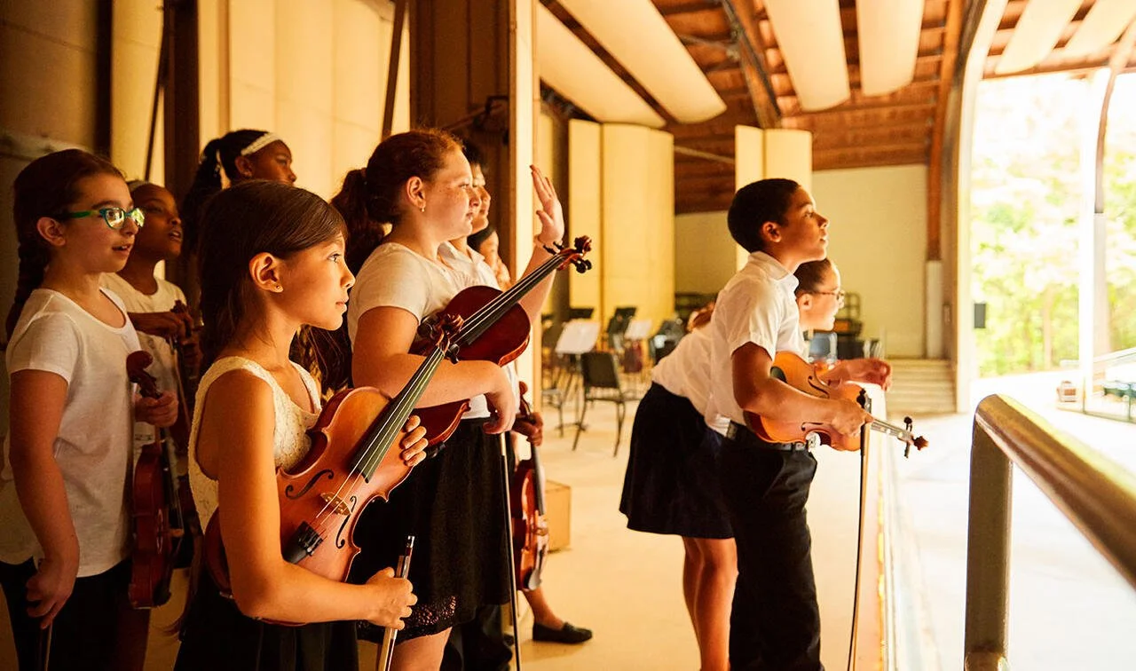 Group of children in a music rehearsal room, some holding violins, watching a performance on stage.