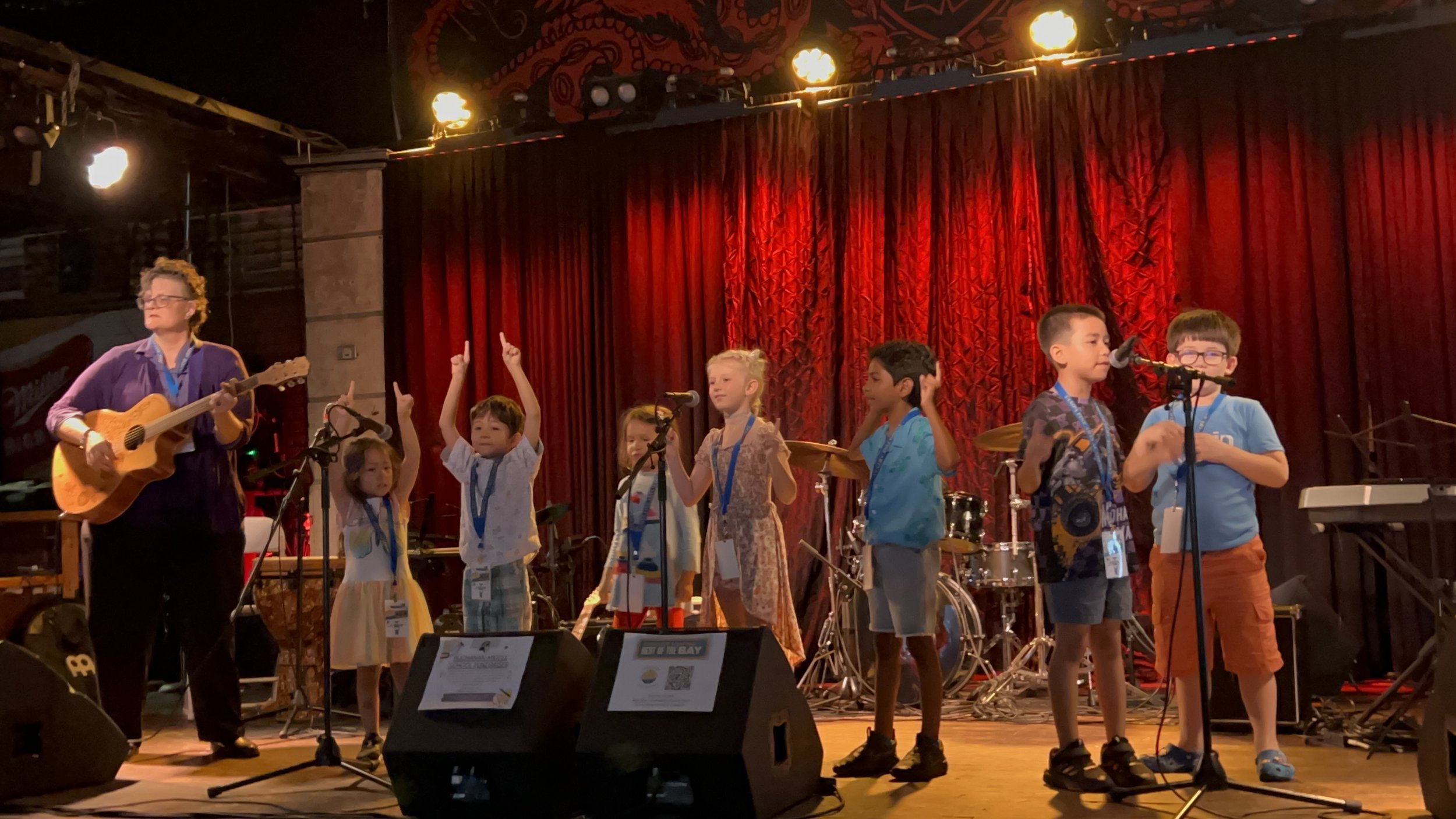 Young children performing on stage with a woman playing guitar. The children are standing in front of a red curtain, some raising their hands or holding microphones, with musical instruments and stage equipment visible.