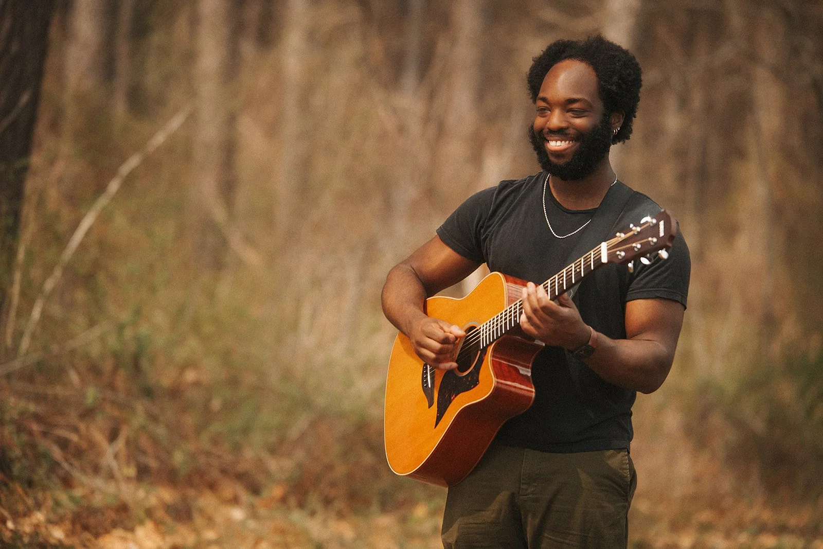 A man with a beard and curly hair smiling while playing an acoustic guitar outdoors in a wooded area during daytime.