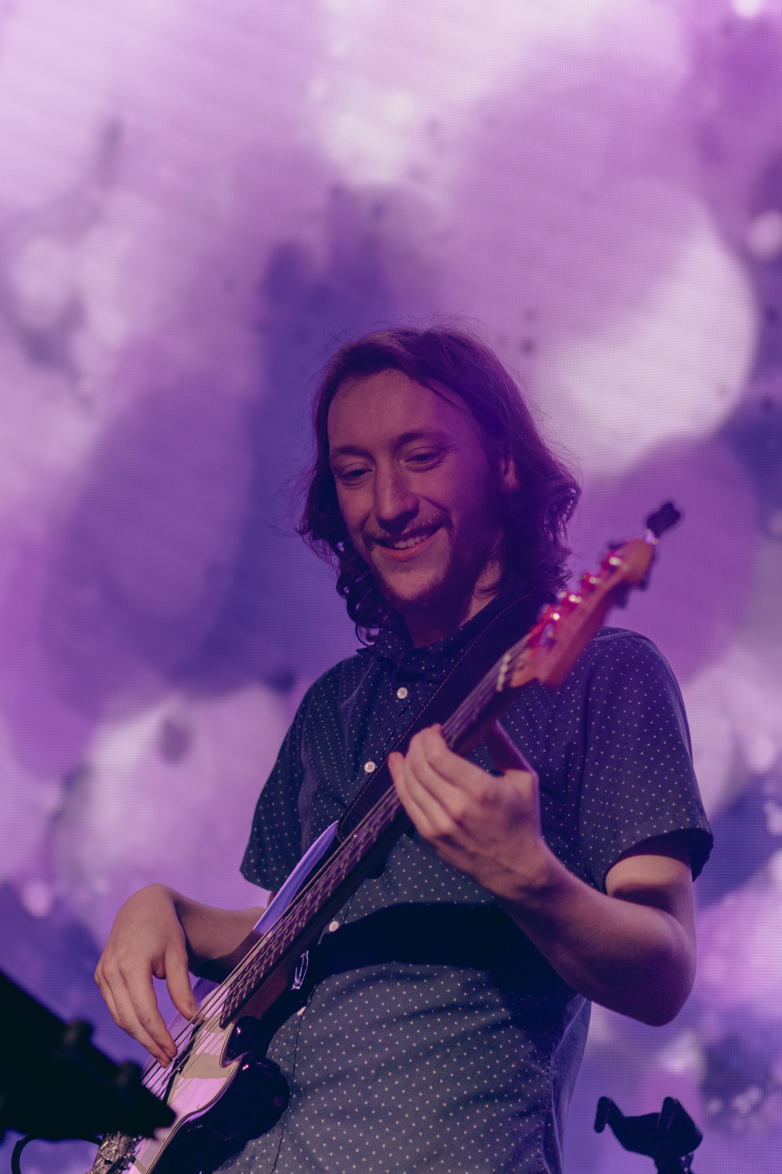 A man with long hair playing an acoustic guitar on stage with a background of large smoke or fog clouds illuminated in purple.