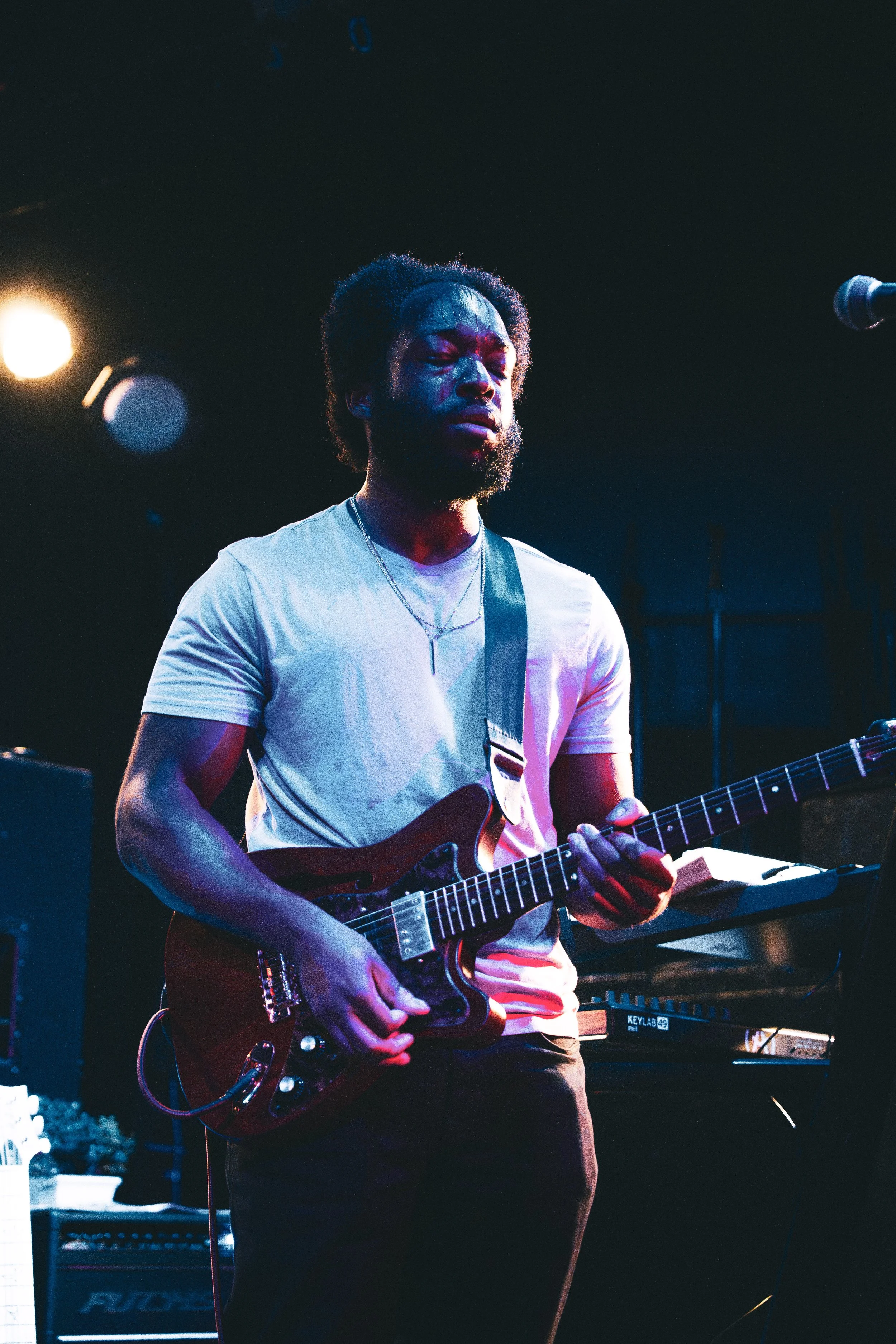 A man in a white t-shirt playing an electric guitar on stage with closed eyes, illuminated by stage lights, with music equipment visible in the background.