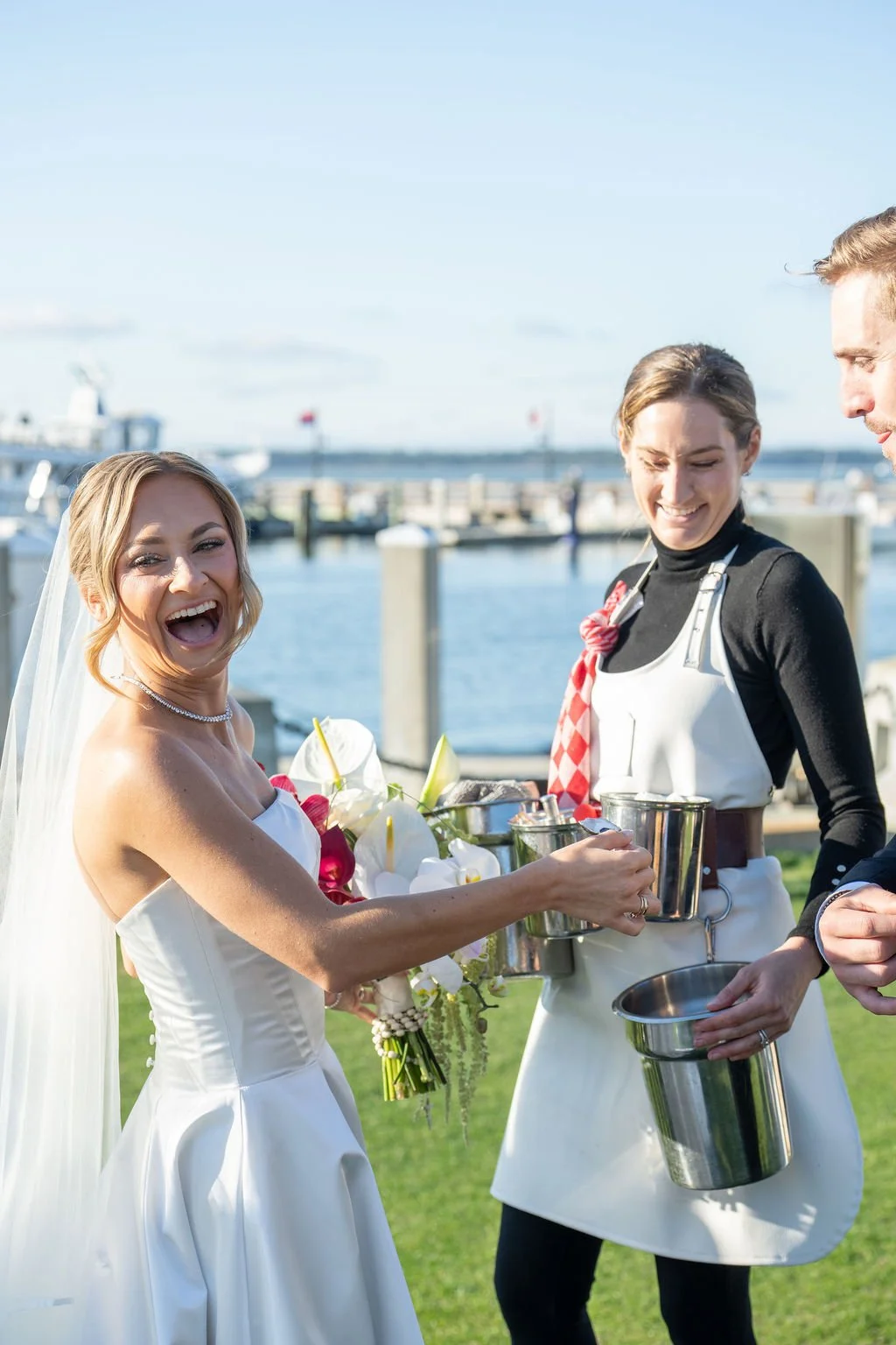 A bride smiling and holding a bouquet during an outdoor wedding near a marina, with a server handing her a drink and guests nearby.