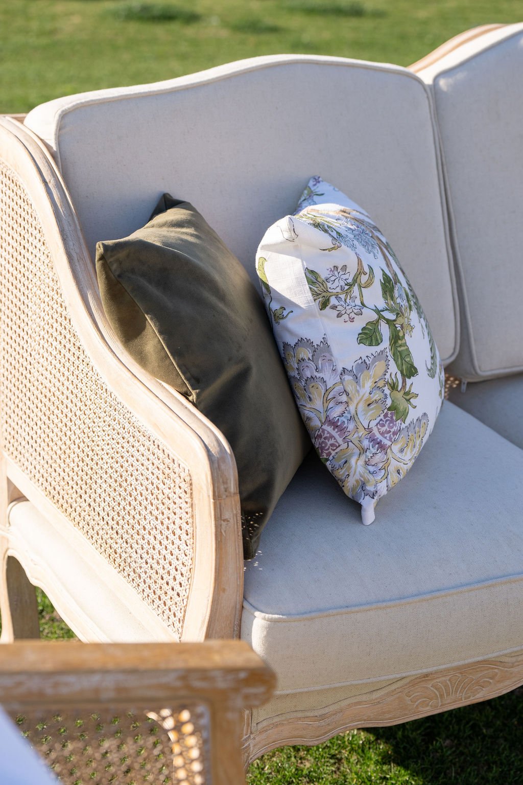 A vintage style cream-colored sofa with a woven rattan backrest, placed outdoors on grass, with two decorative pillows: one dark green and one floral with shades of purple, yellow, and green.