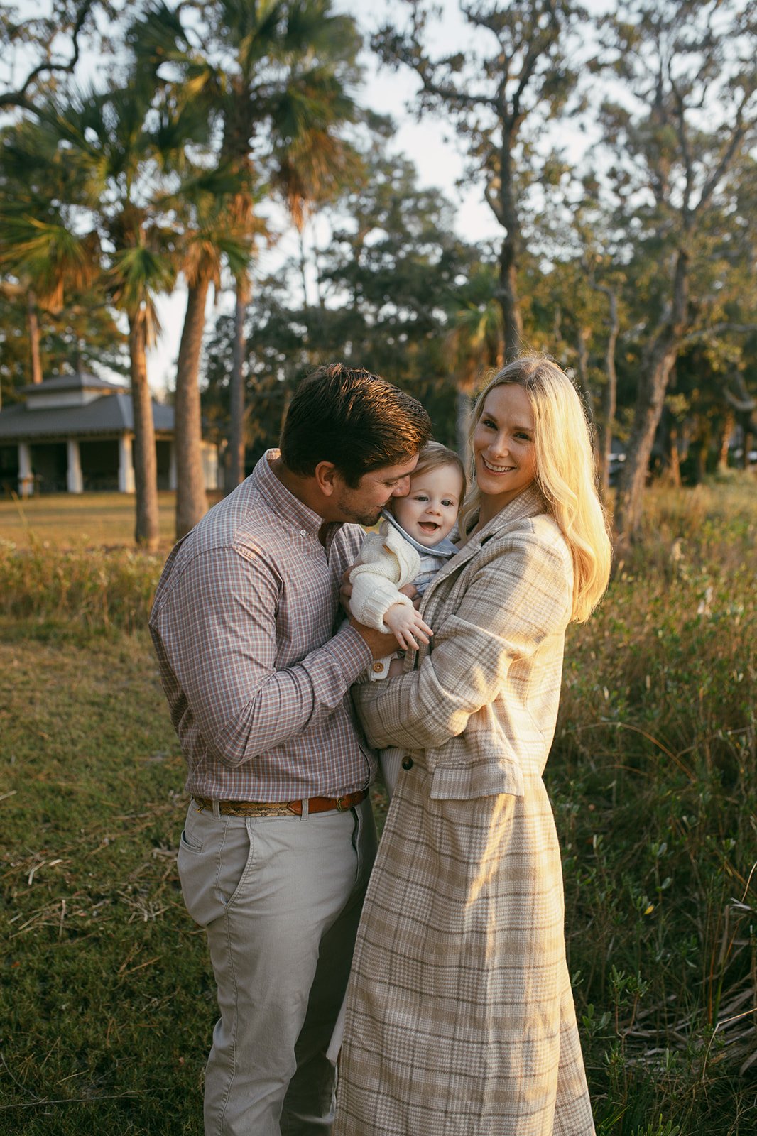 A family of three outdoors, with the father kissing the child's cheek, the mother holding the smiling child, in a park or garden setting during sunset.
