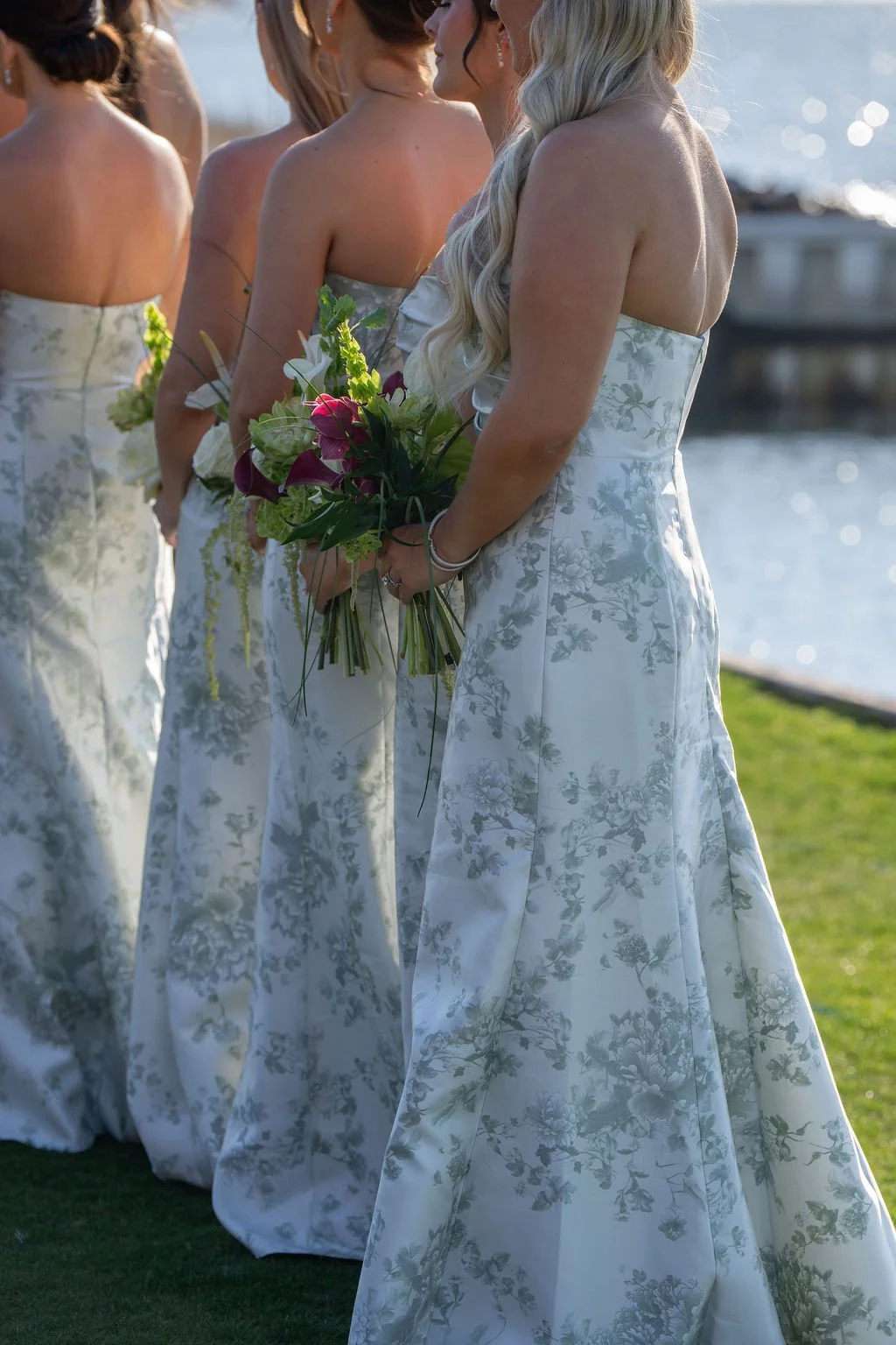 A line of women dressed in white floral strapless dresses, holding bouquets, standing outdoors near a body of water.