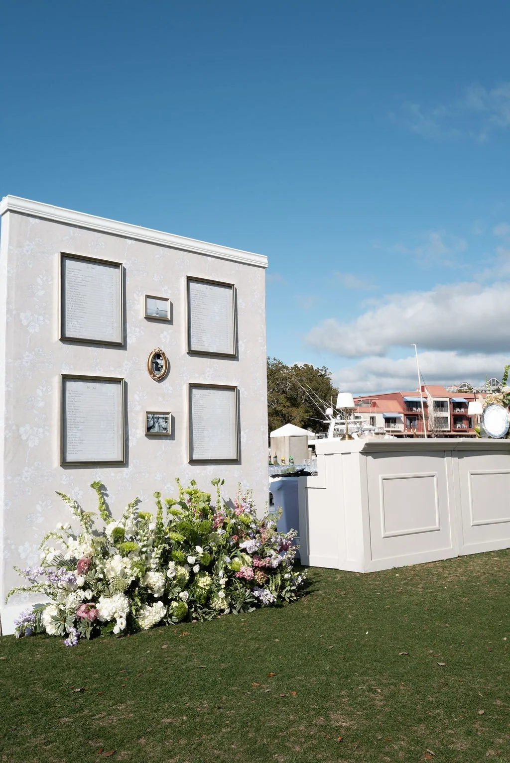 A memorial wall with framed names and flowers at its base, located outdoors near a marina with boats and modern buildings in the background under a blue sky.