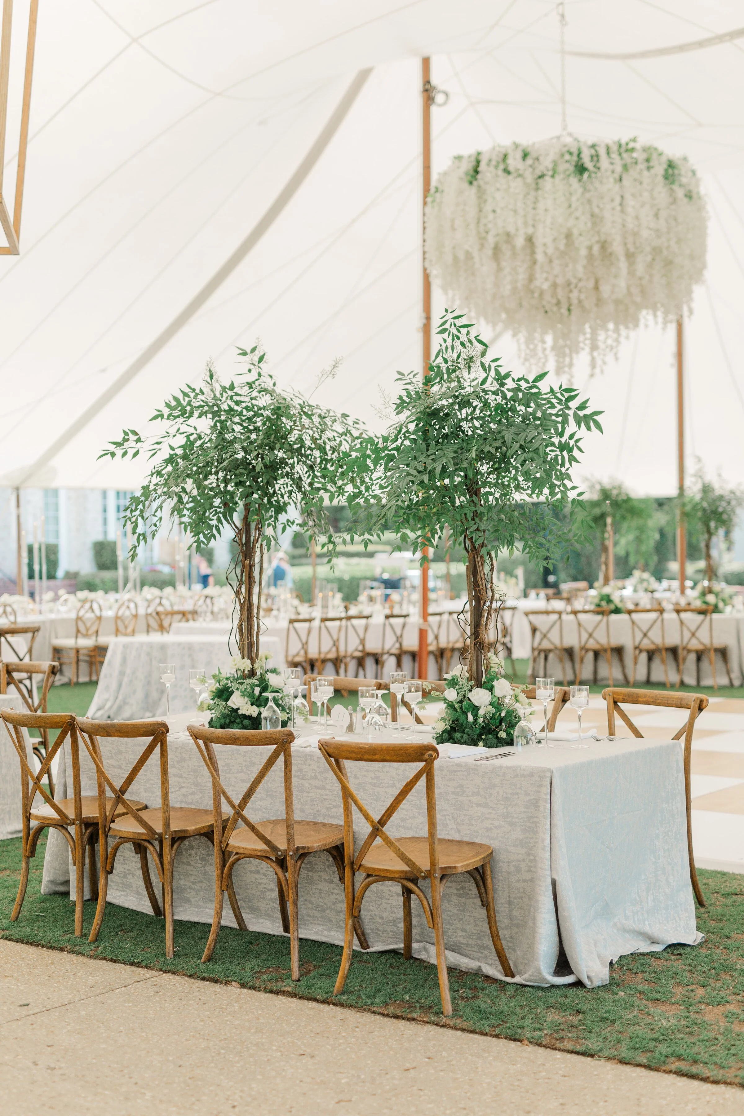 Elegant tables set with white tablecloths, glassware, and floral arrangements, under a large white canopy with hanging floral decorations and potted trees, at an outdoor event space.