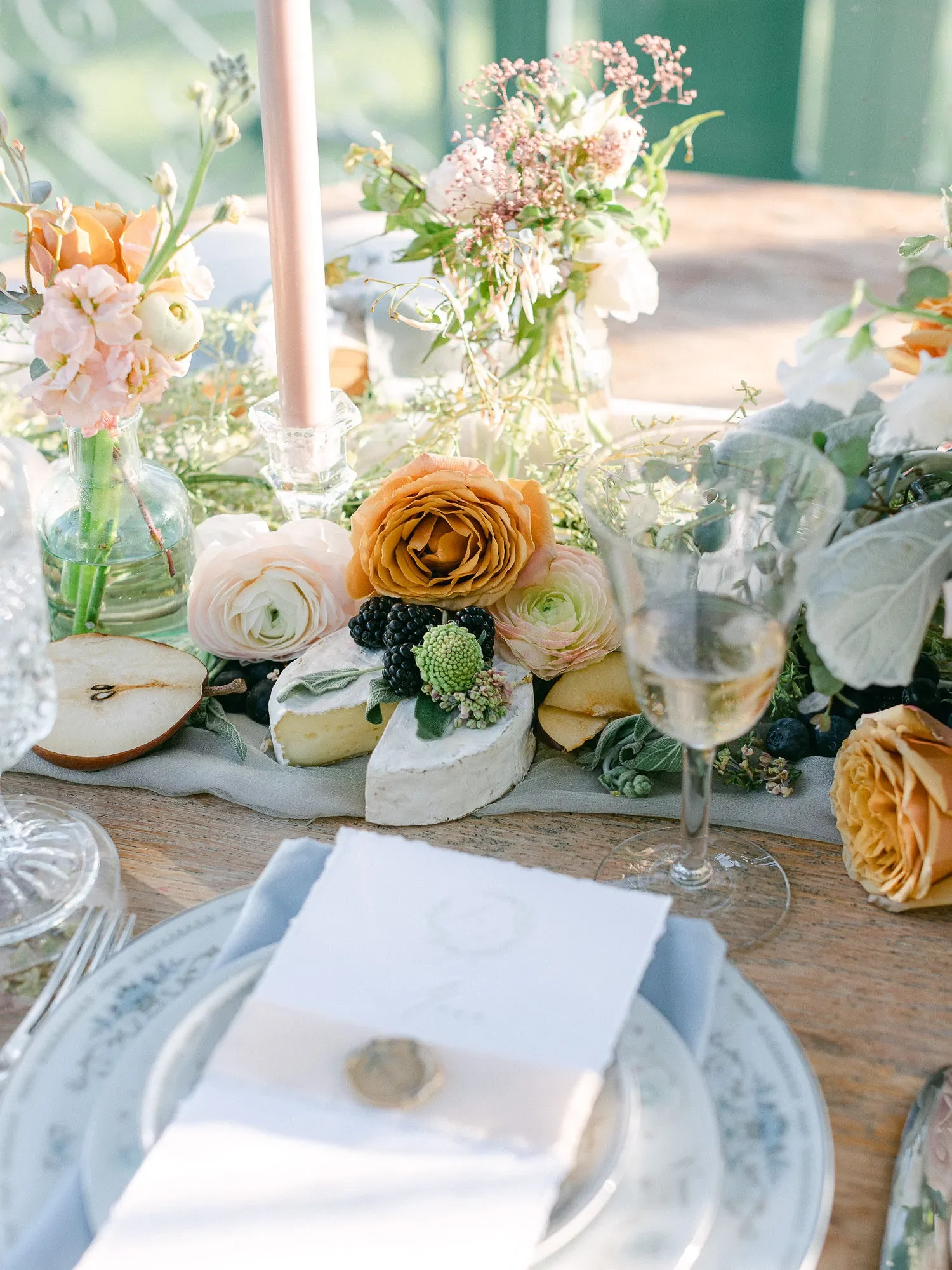 A table setting with flowers, cheese, blackberries, and a birthday cake with a candle in a clear glass container. The focus is on the cake with a white napkin and a plate with silverware in the foreground.