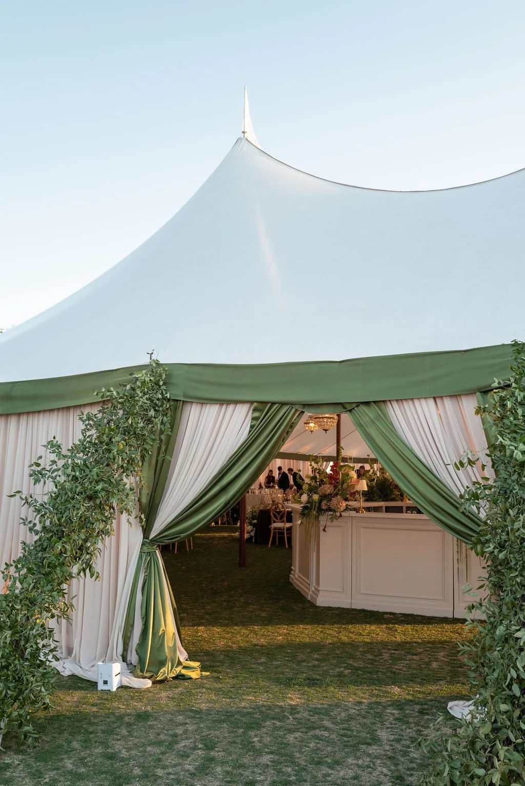 Entrance of elegant outdoor event tent with green and white drapes, greenery, and visible interior with people and floral decor.