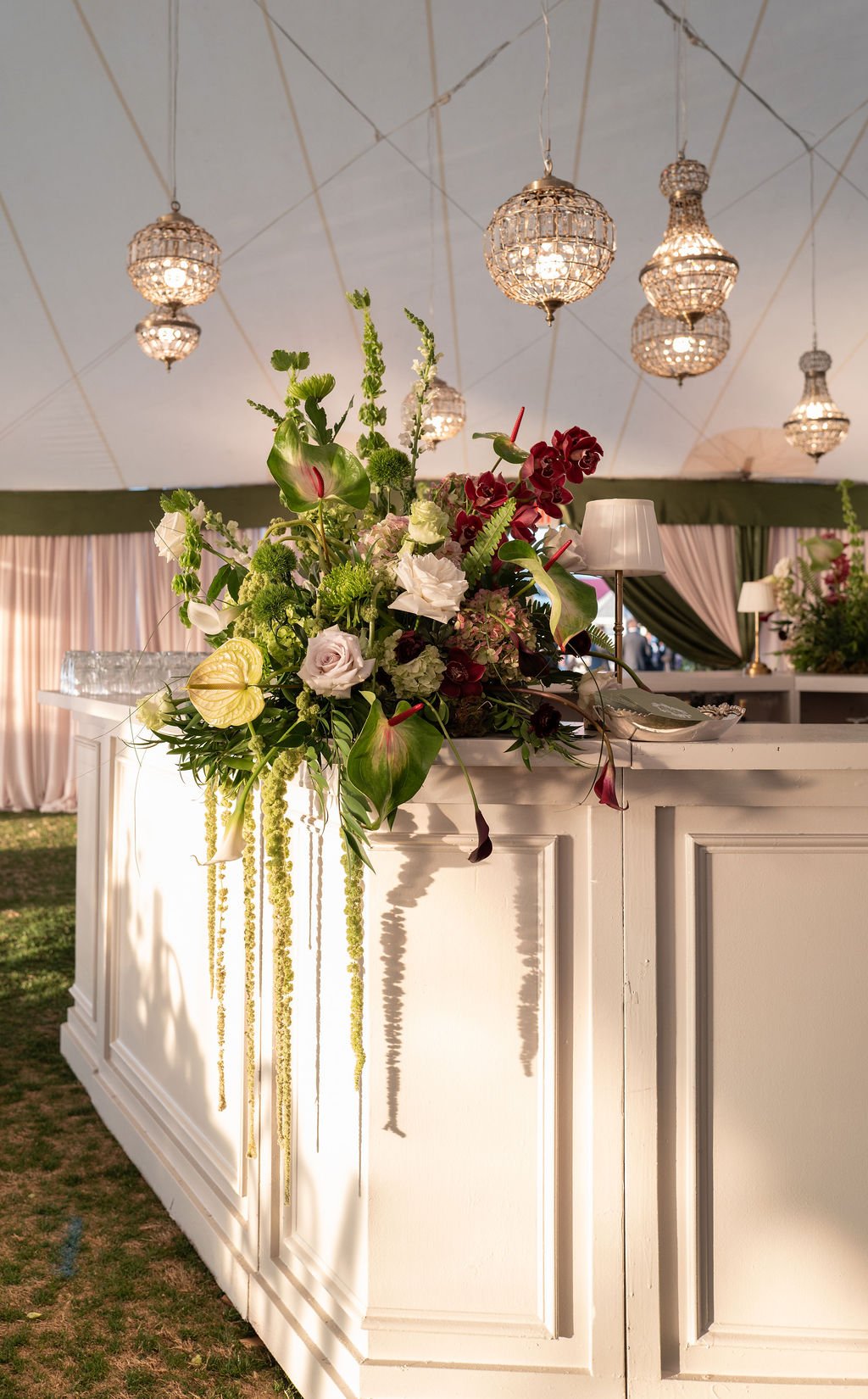 Elegant floral arrangement on a white bar counter with hanging crystal chandeliers and draped pink and green curtains in a decorated event space.