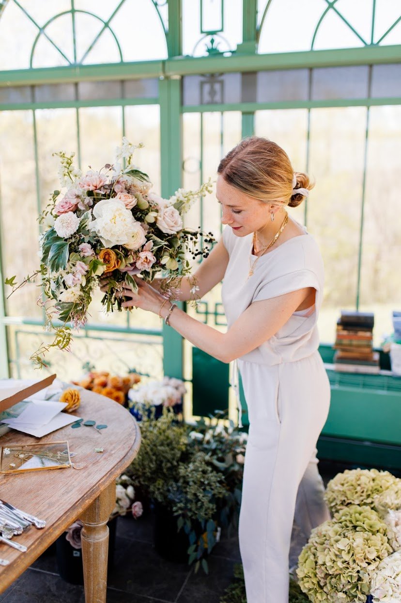 A woman arranging a large bouquet of pink, white, and green flowers in a greenhouse.