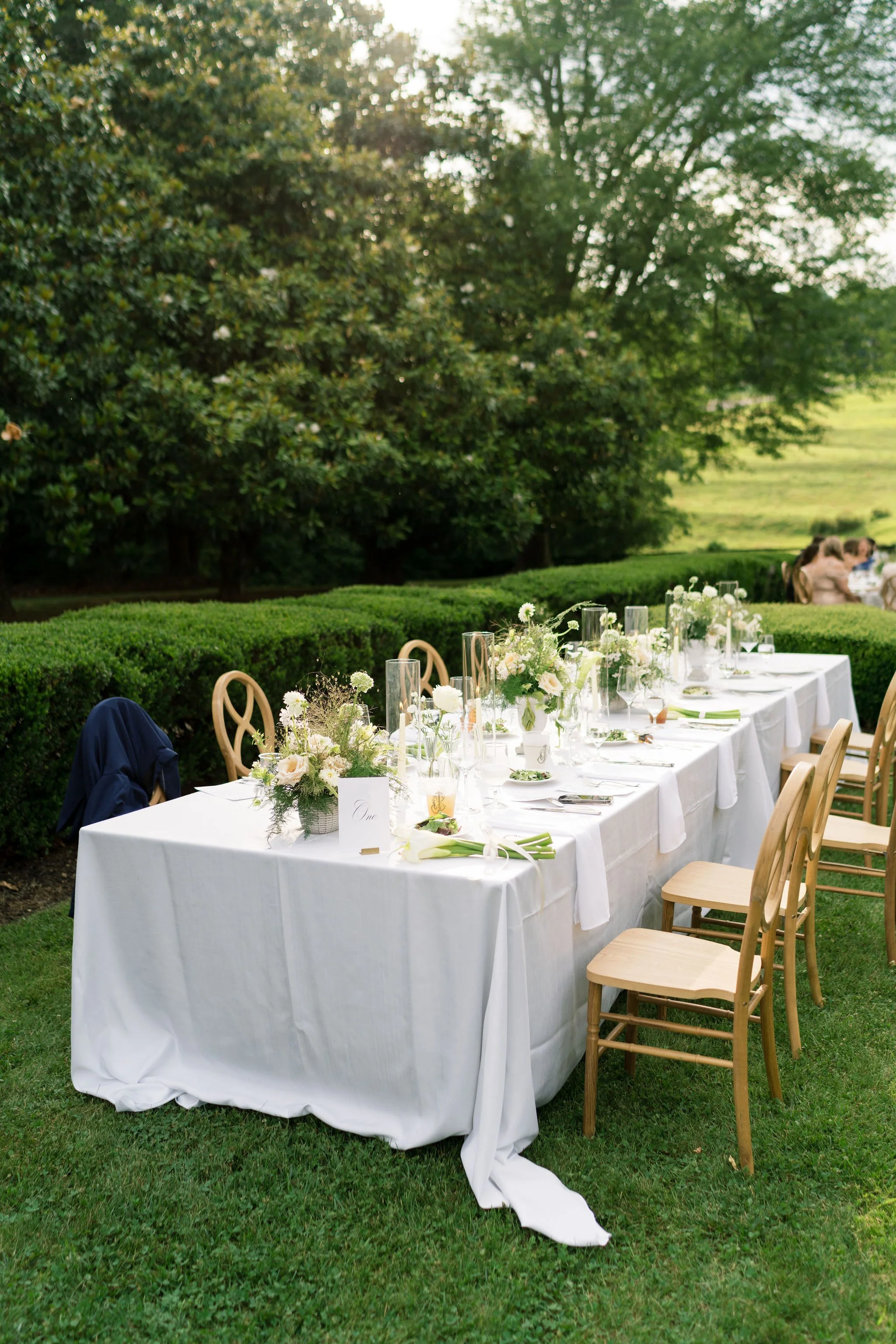 Outdoor dining setup with a long white tablecloth, floral centerpieces, glassware, and chairs, set in a lush green garden with trees and grass.