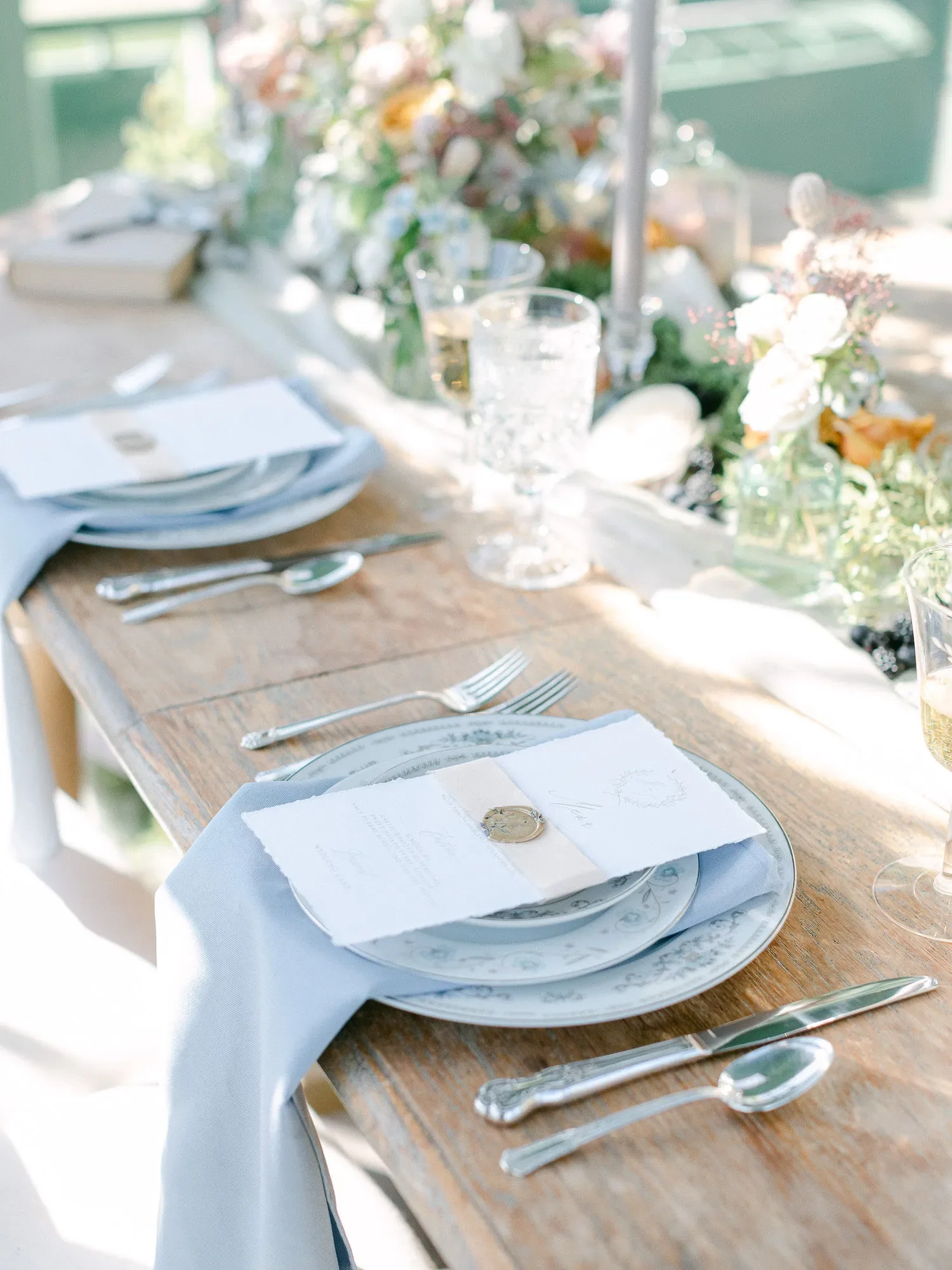 Elegant table setting with white and blue china, silver cutlery, a white napkin, and a menu card, arranged outdoors with floral decorations and glassware.