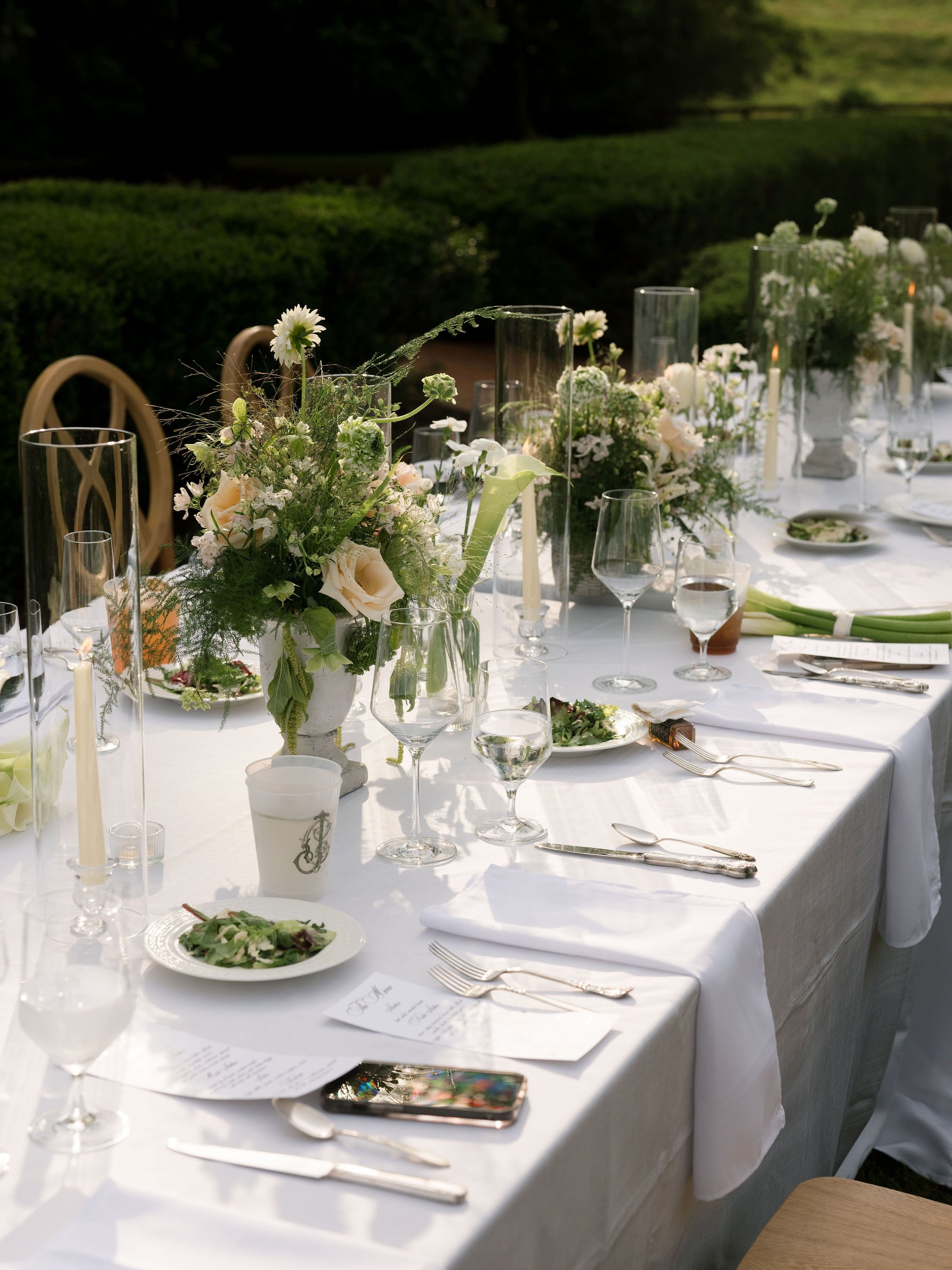 A long outdoor dining table decorated with floral centerpieces, candles, wine glasses, plates, and silverware, set for a meal in a garden or park setting.