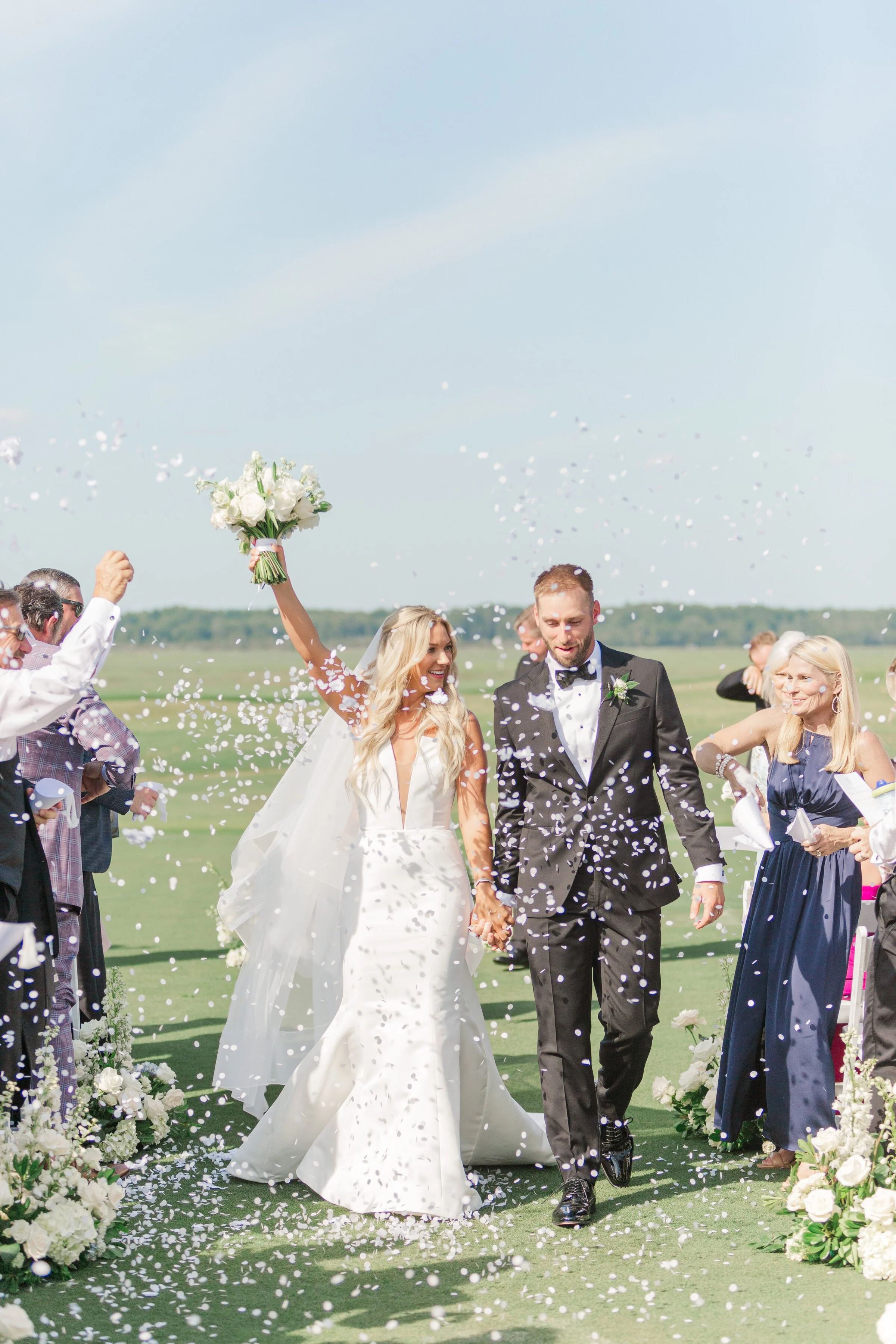 A newlywed couple holding hands walking through a shower of white flower petals at an outdoor wedding ceremony. The bride is holding a bouquet of white flowers, and both are dressed in formal wedding attire with guests on either side.