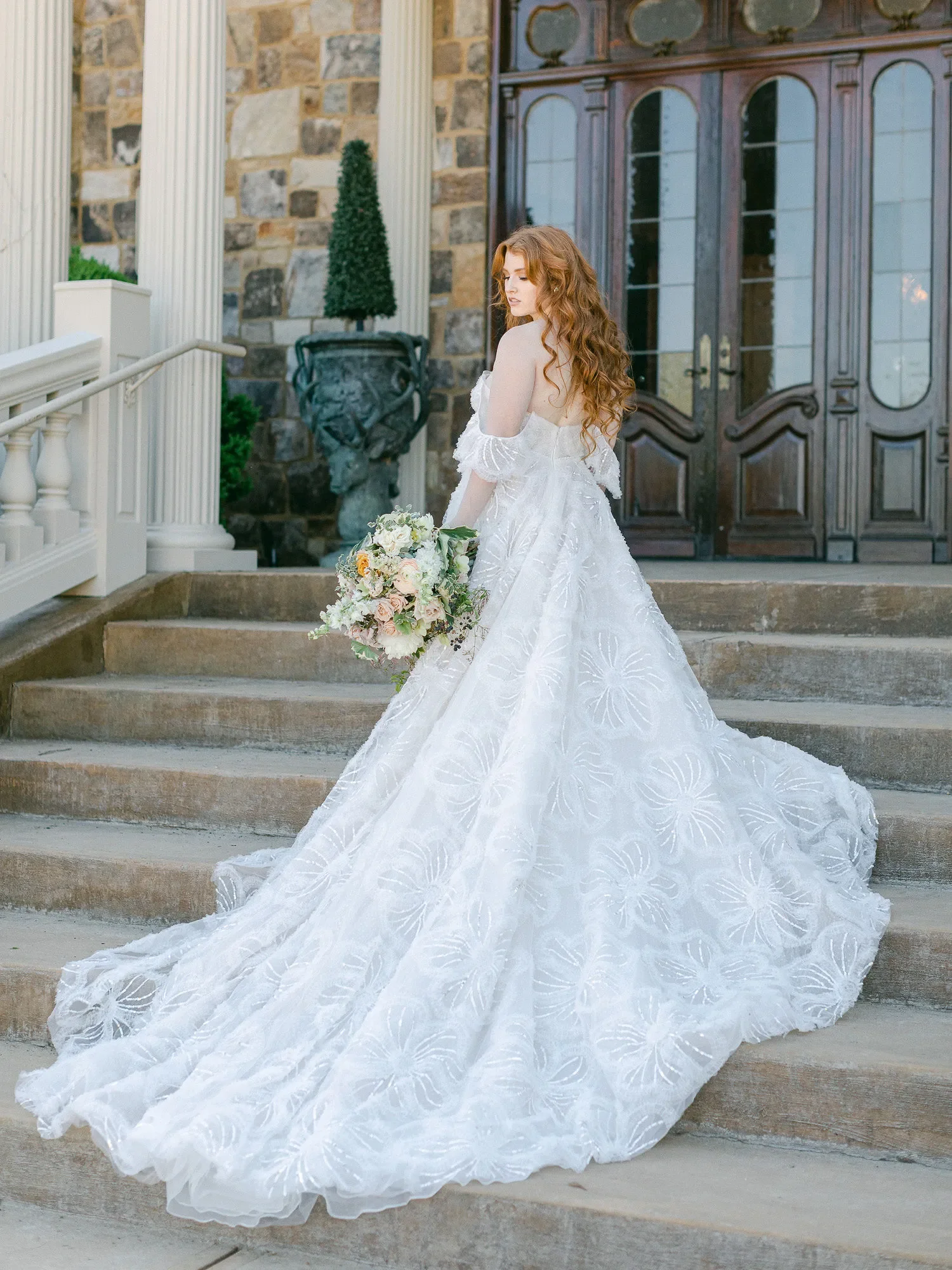 A young woman with red curly hair in a wedding dress holding a bouquet of flowers on stone steps in front of a building with large wooden doors and stone walls.