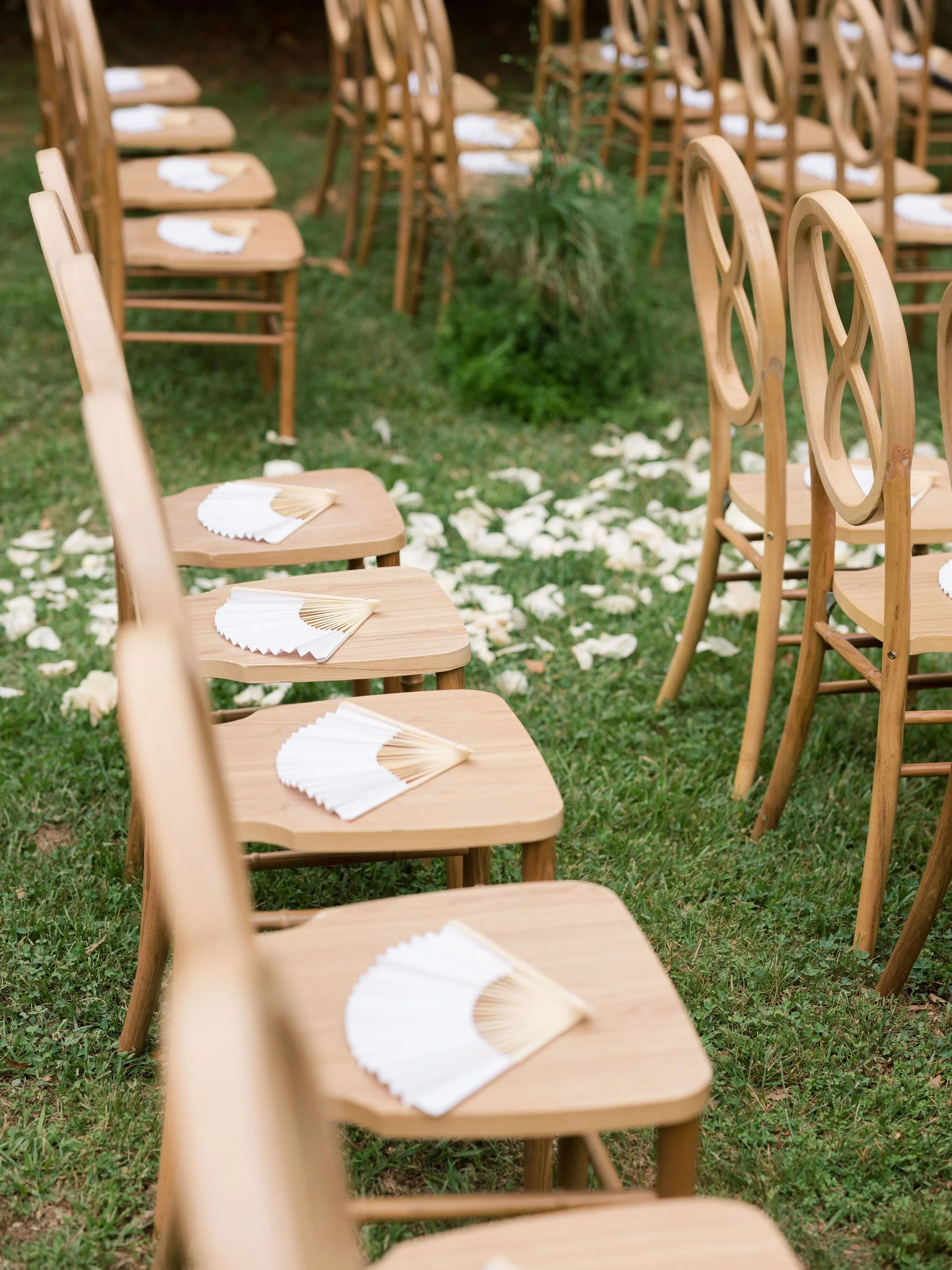 Wooden chairs with white cloth napkins and paper fans on a grassy outdoor area, arranged for a gathering or ceremony.
