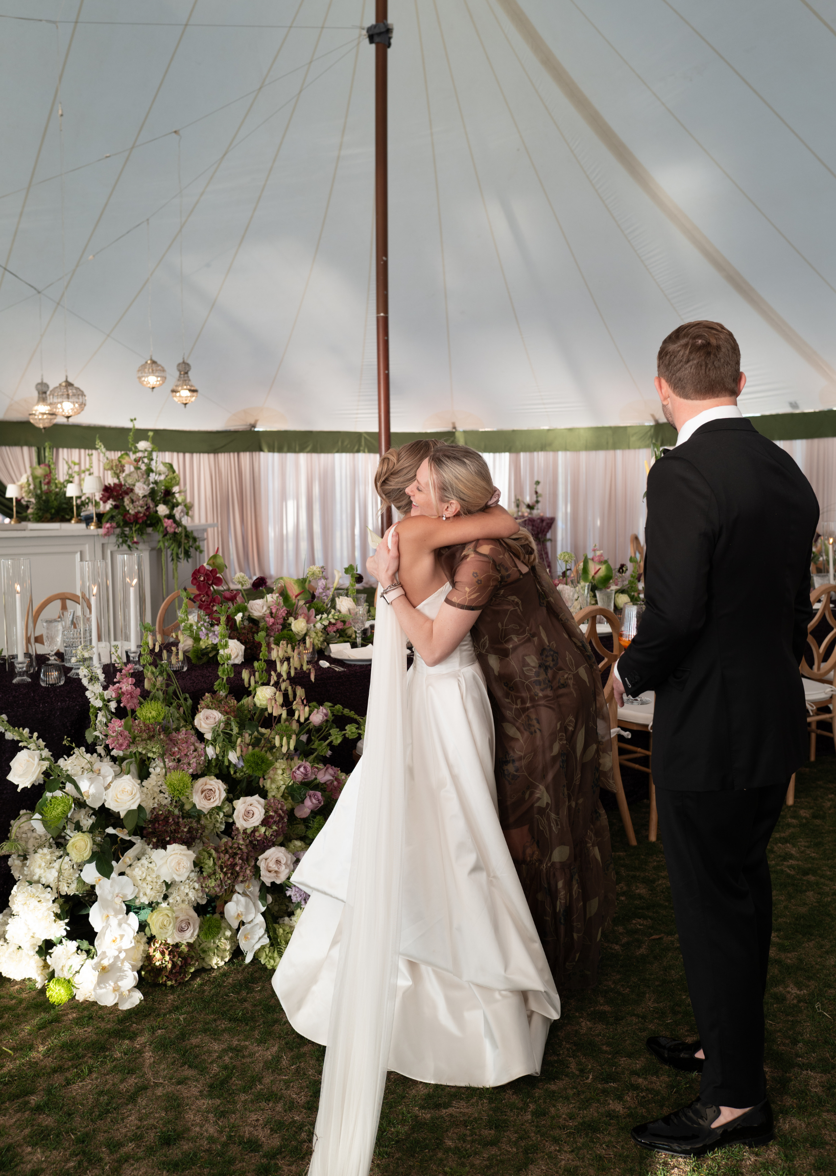 A bride embraces a woman in a brown dress at a wedding reception with floral decorations and a large white tent.