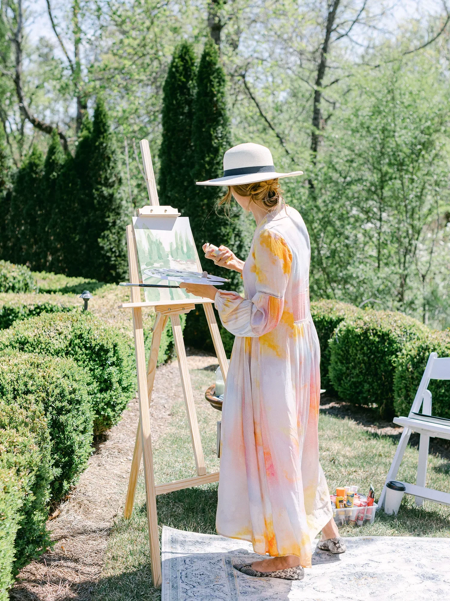 Woman painting outdoors on an easel, wearing a wide-brimmed hat and a colorful long dress, with art supplies and a water bottle nearby, surrounded by greenery and bushes.