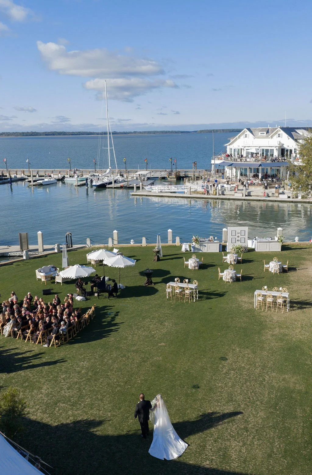 Wedding ceremony setup outdoors on a grassy area near a waterfront with boats and a seaside building, with guests seated and a bride and groom walking.