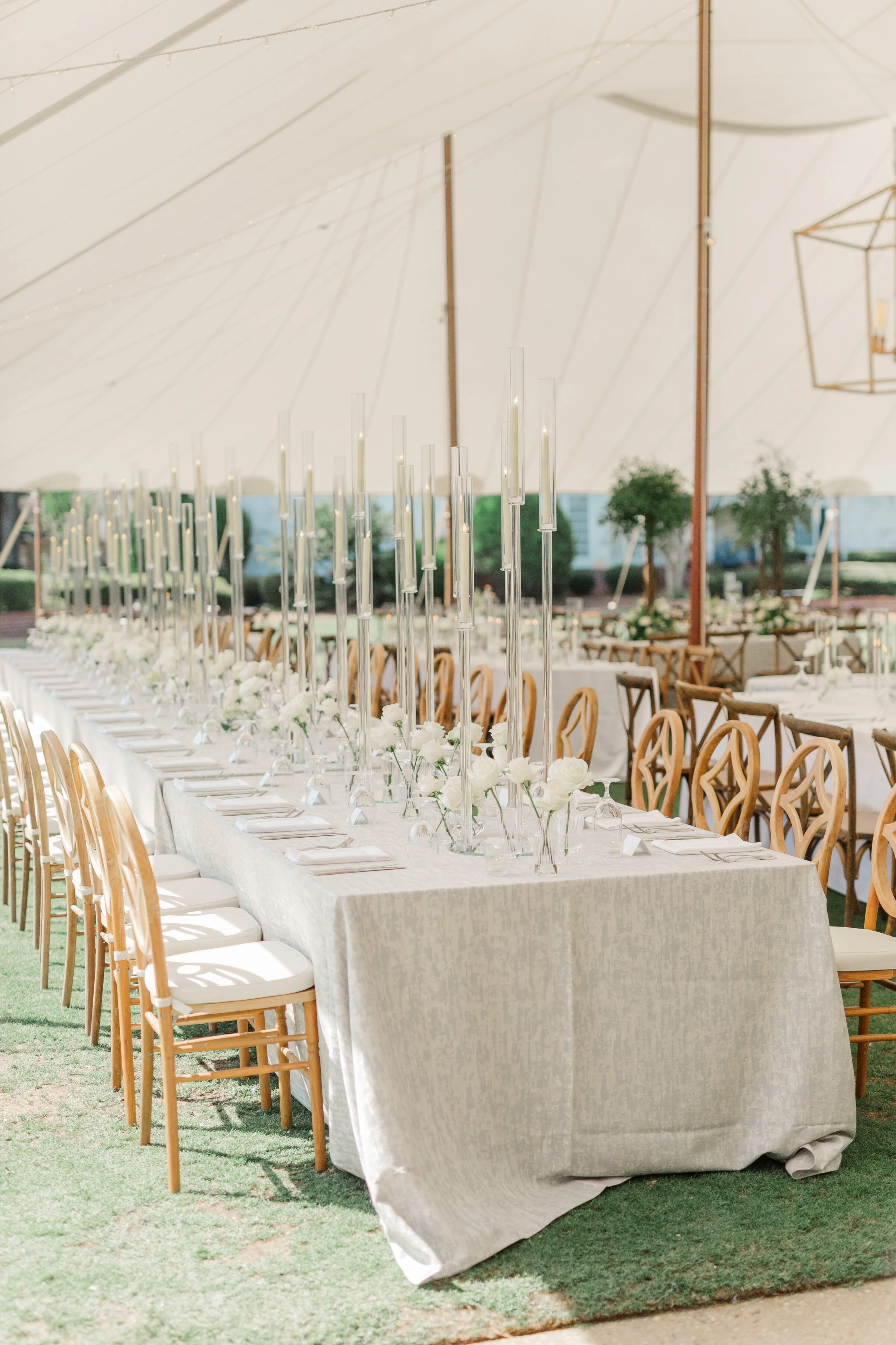 Long rectangular table set for a formal event with white tablecloth, floral centerpieces, tall clear candle holders, and wooden chairs in an outdoor tented area with grass and trees outside.