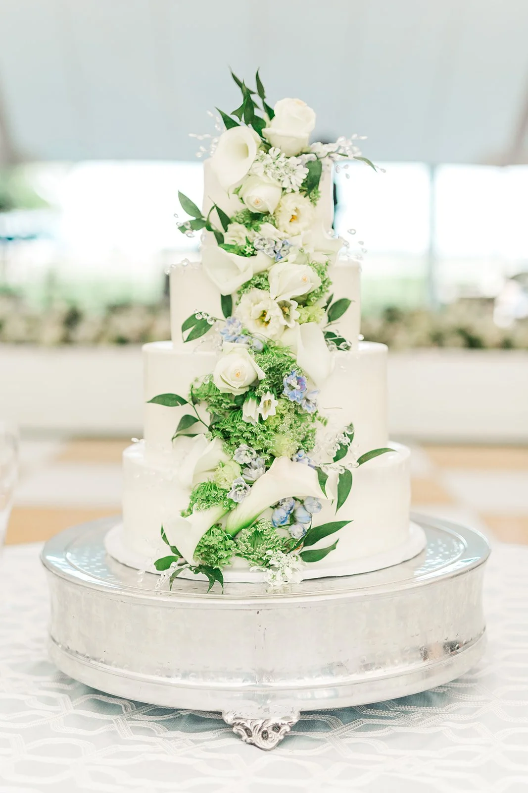 A tiered white wedding cake decorated with white and light purple flowers and green foliage, placed on a silver cake stand.