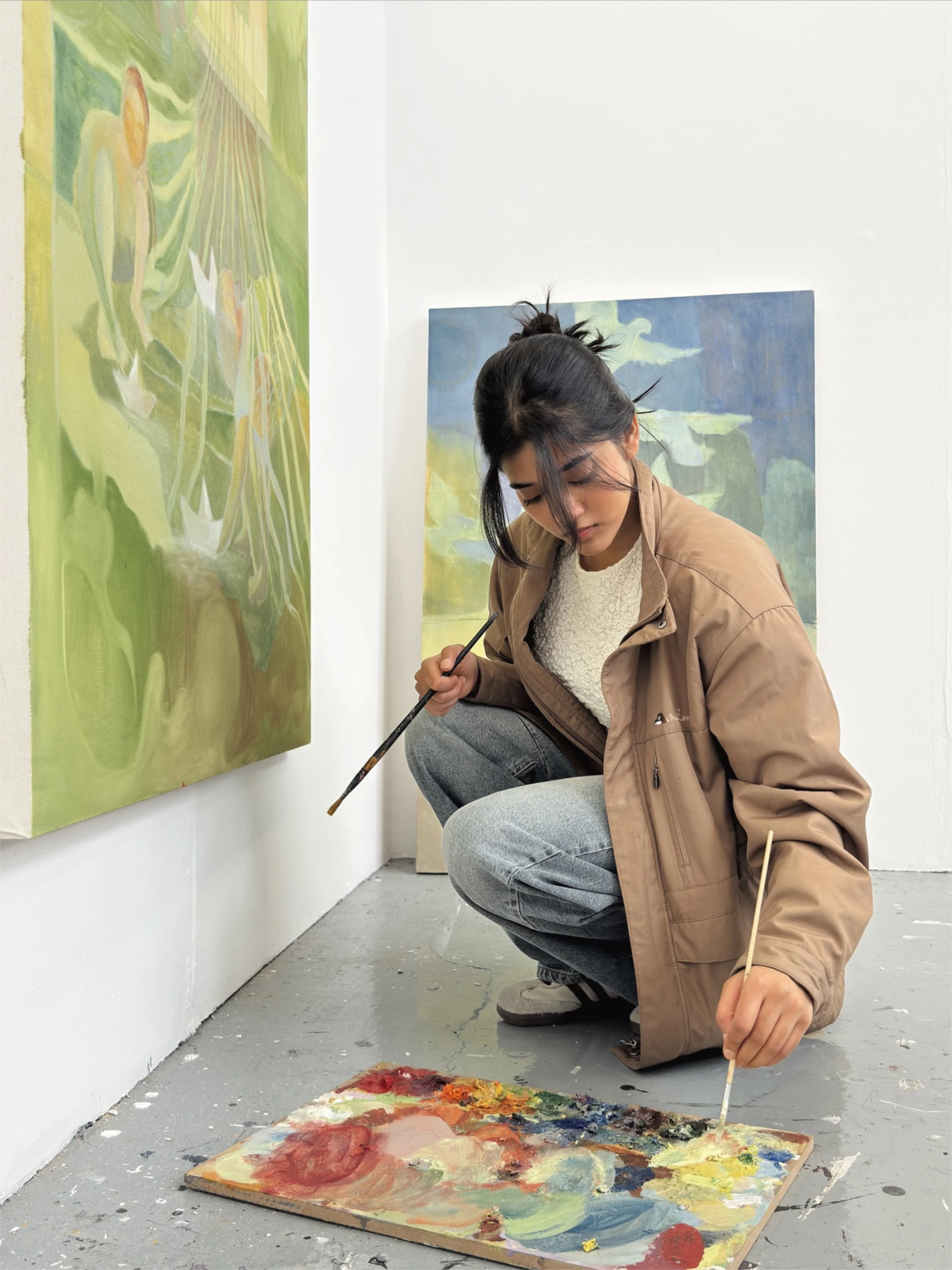 A woman crouching on the floor of an art gallery, painting on a large canvas with bold, colorful strokes. She is wearing a beige jacket and gray jeans, with her black hair tied up in a messy bun. There are paintings on the gallery white walls, and the floor is a gray concrete with some paint splatters.