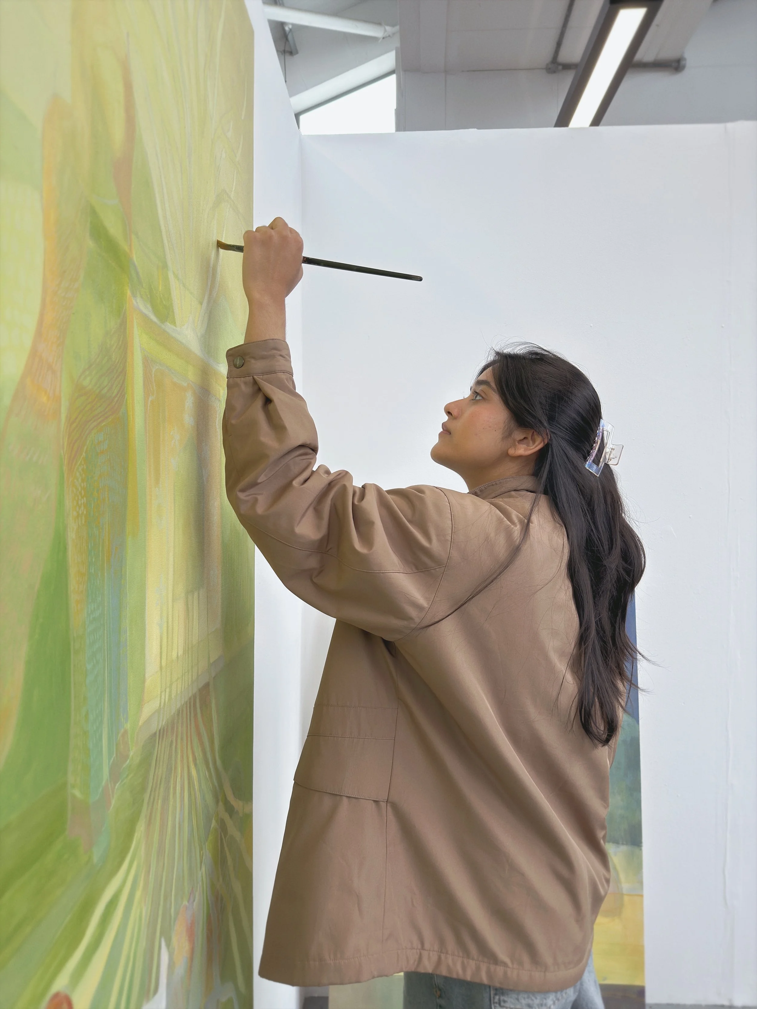 A woman with long dark hair, wearing a light brown jacket and jeans, is painting a colorful mural on a wall in an art studio. She is holding a paintbrush and focused on her artwork.