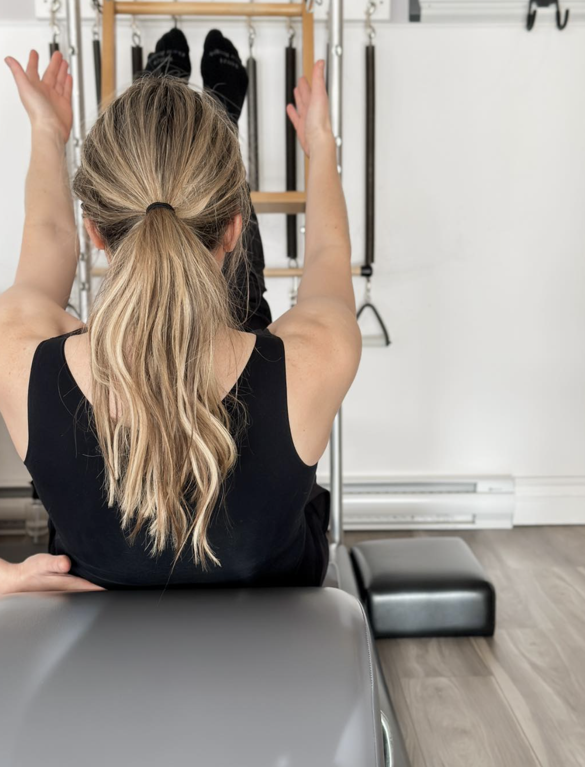 A woman with blonde hair tied in a ponytail sitting on a chair, raising her arms while being assisted with physical therapy or exercise in a fitness or rehab facility.