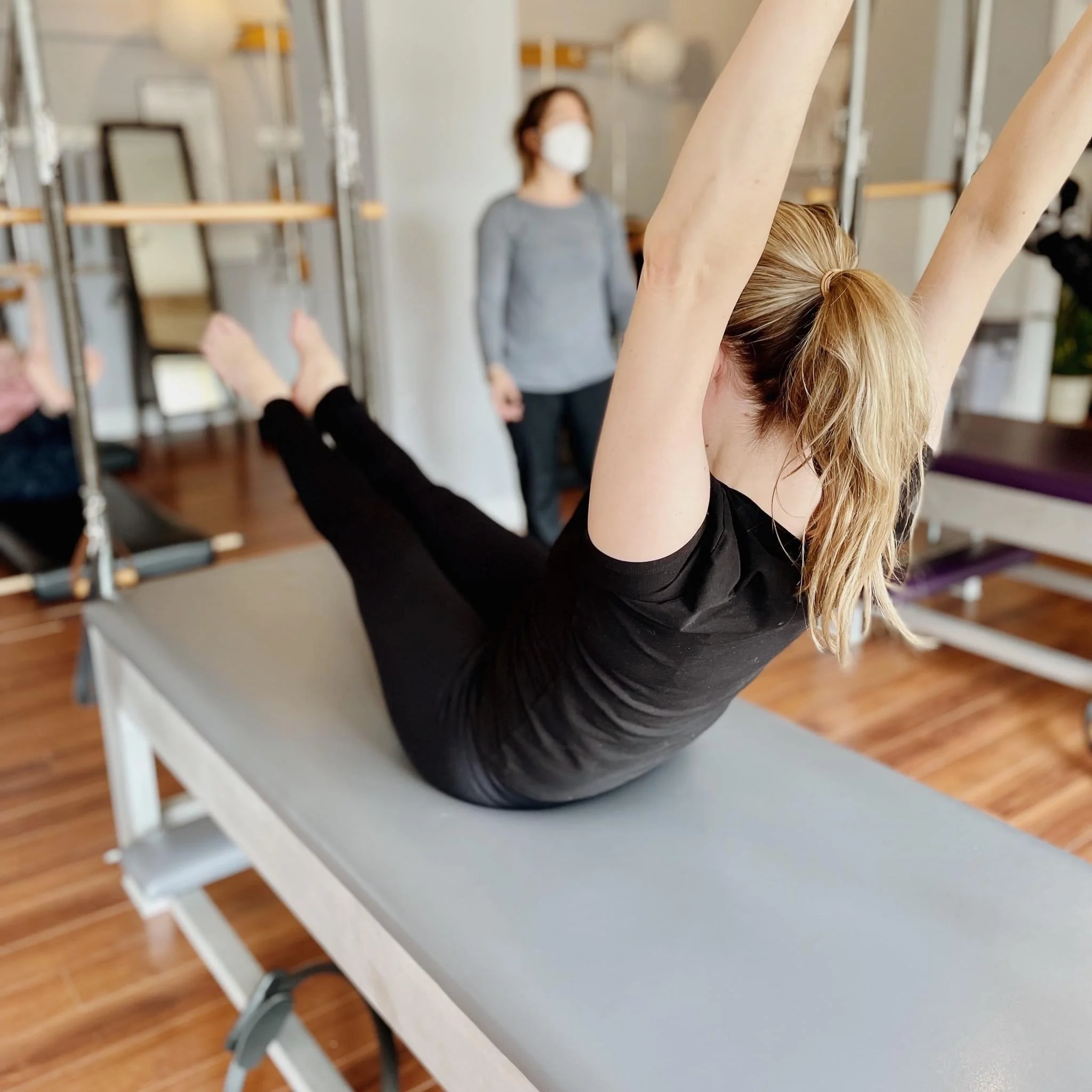 Person with a ponytail doing a Pilates Teaser on a Cadillac, with a teacher and other students in the background.