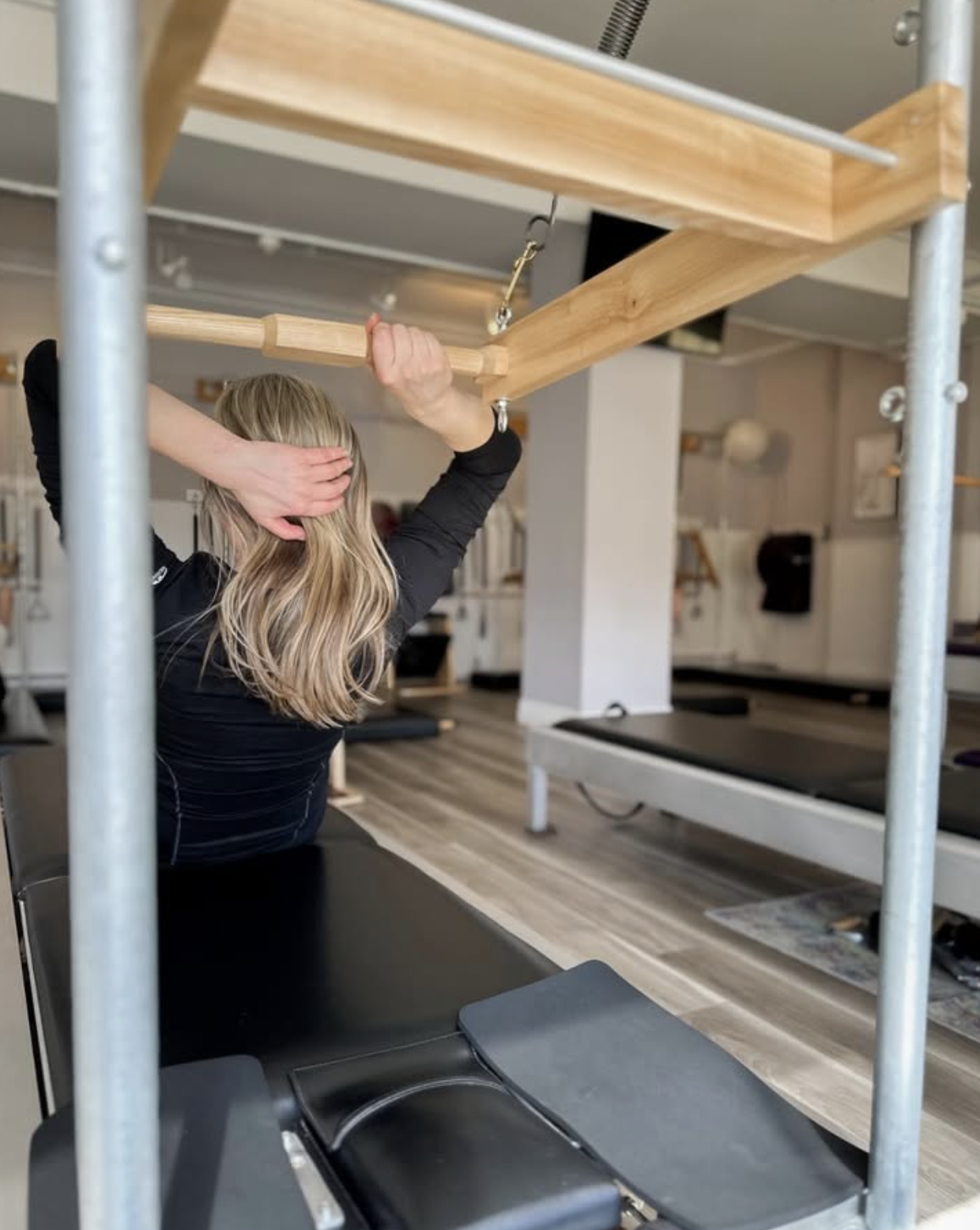A woman with blonde hair doing an exercise on a black workout bench in a gym, with a wooden frame and metal scaffolding around her.