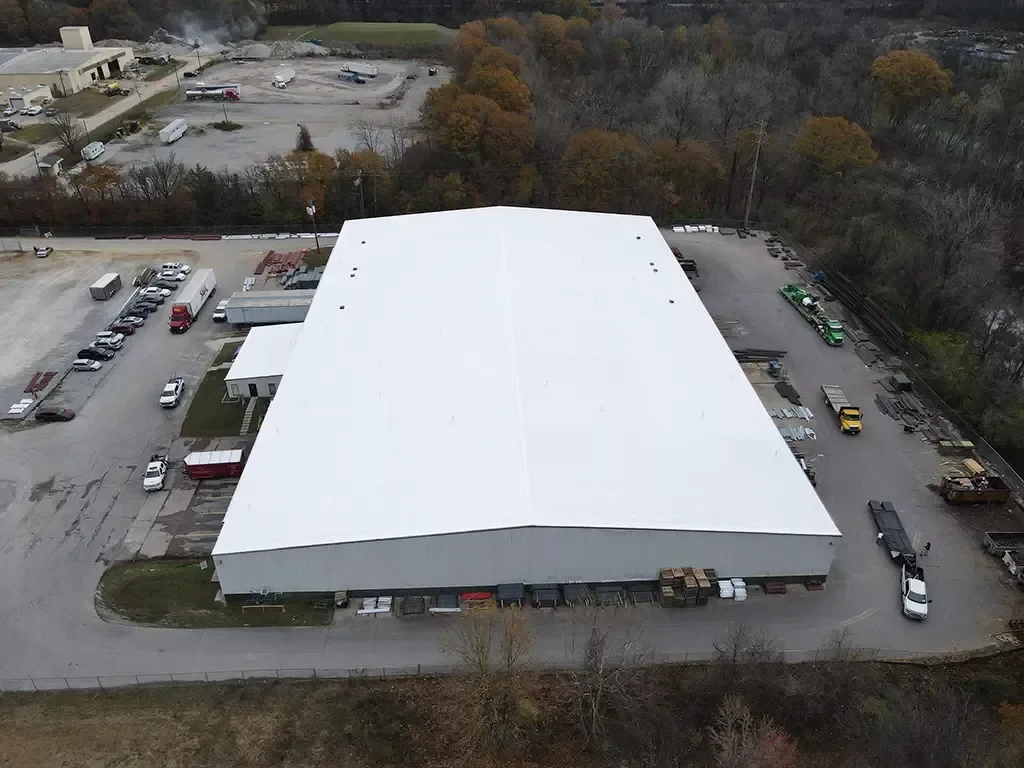 A large industrial warehouse with a white roof, surrounded by parking lots with various vehicles, and wooded area in the background.