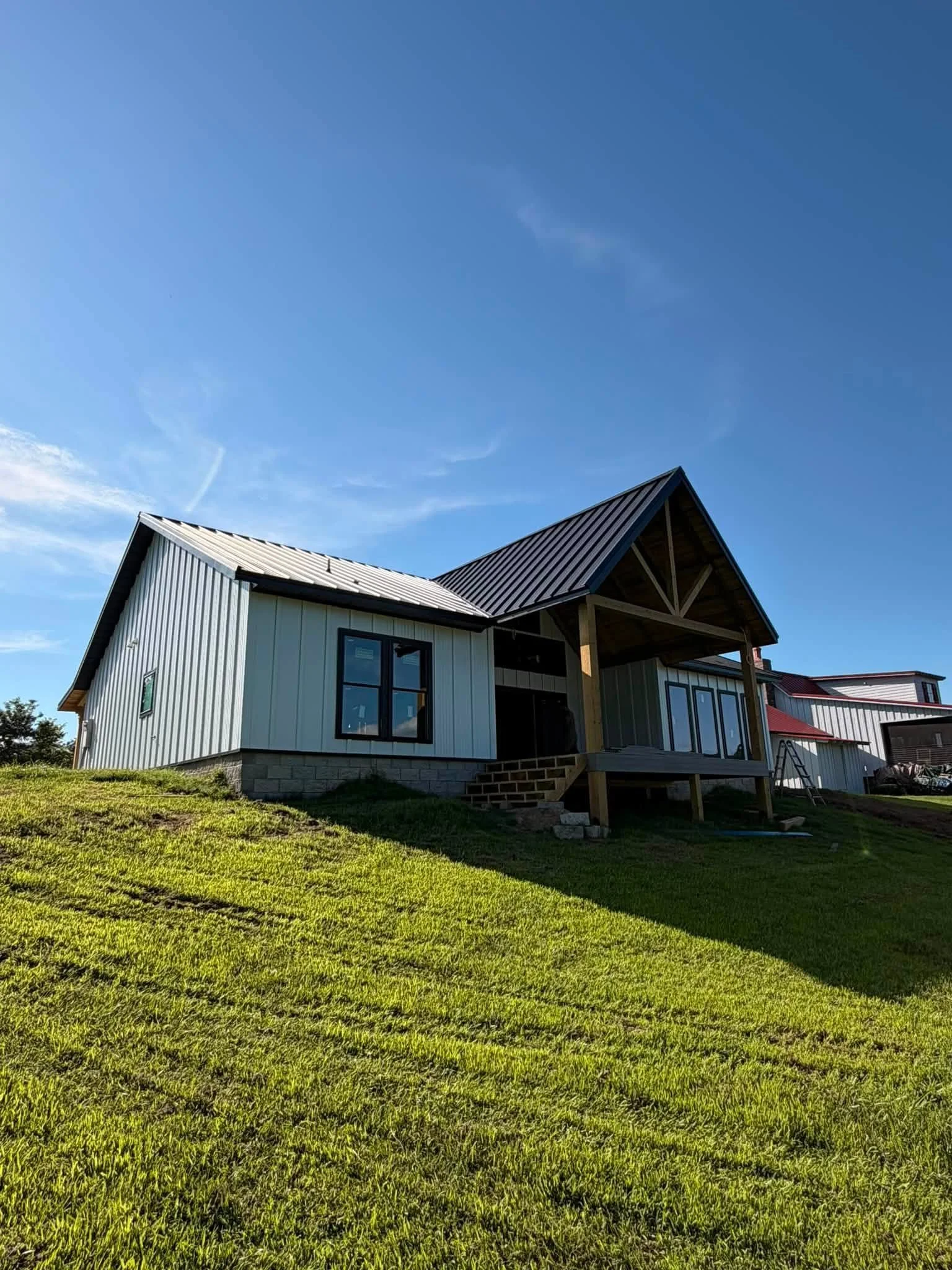 A house with white exterior walls, black window frames, and a metal roof, under a blue sky with some clouds, situated on a green grassy lawn.