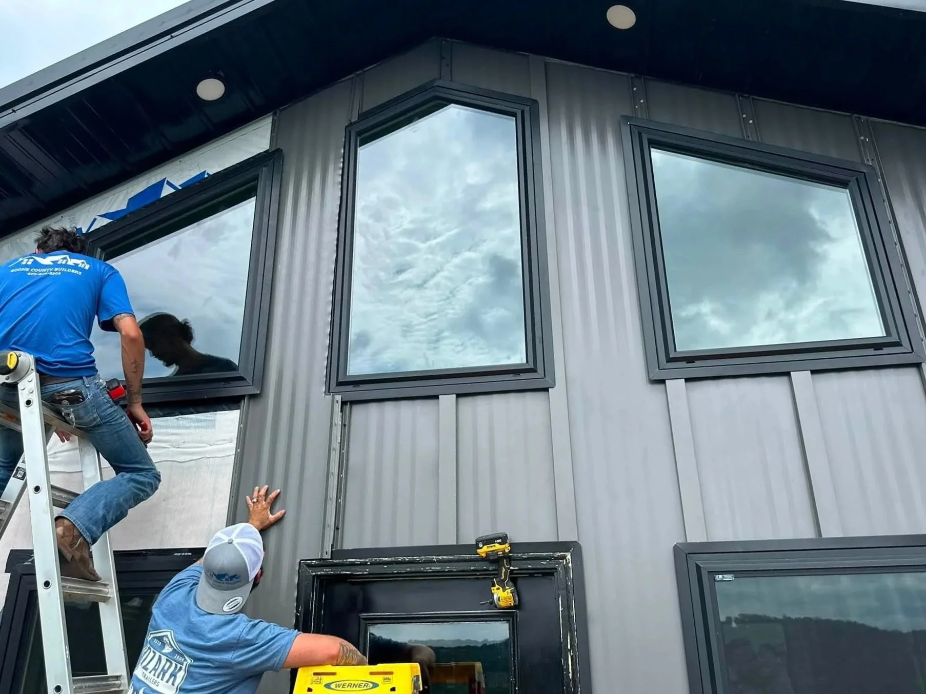 Workers installing or repairing large windows on the exterior of a metal building, with clouds reflected in the glass.